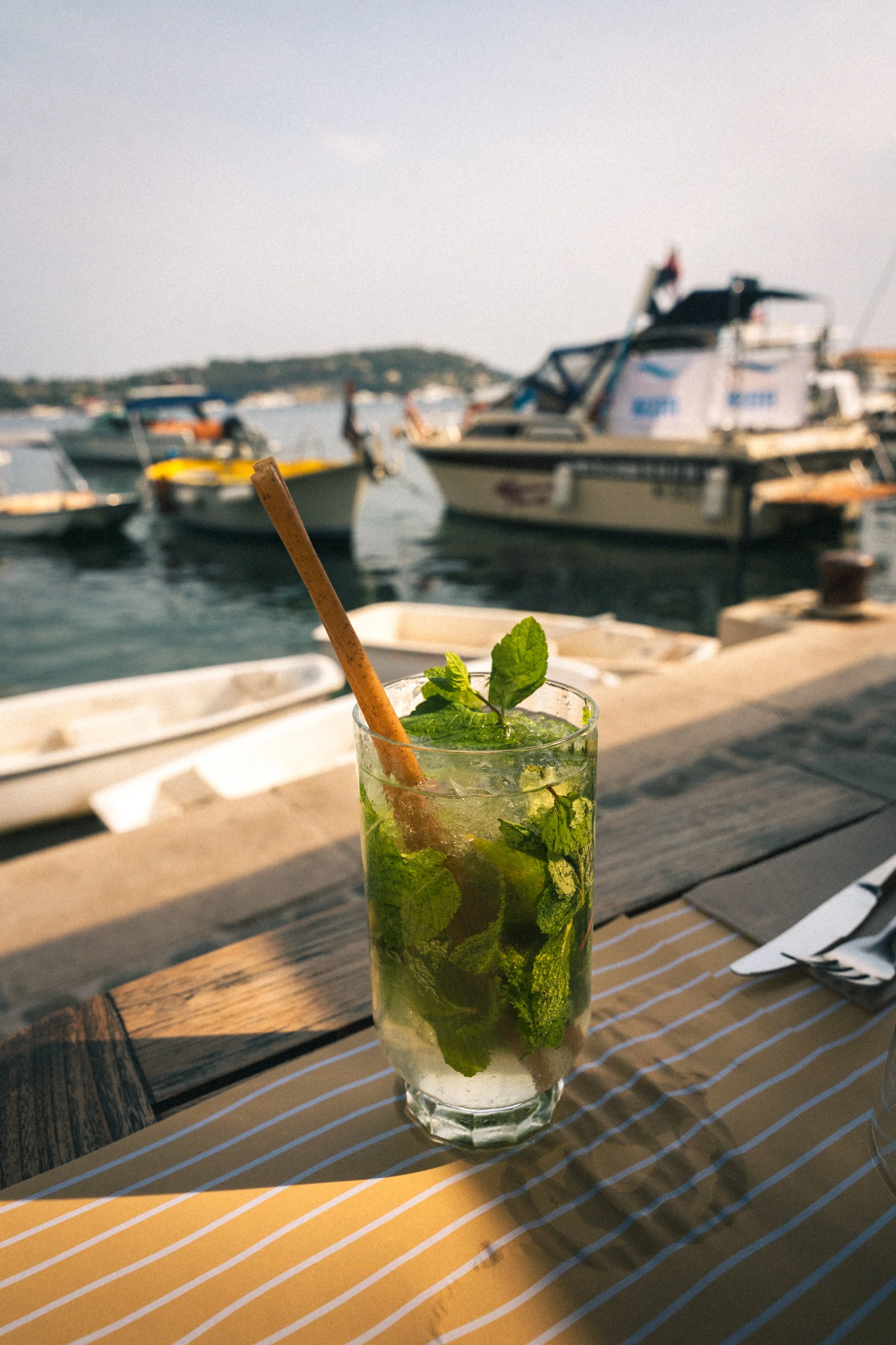 A mojito cocktail with mint leaves, lime, and a brown straw on a wooden table outside with boats docked in the water in the background.
