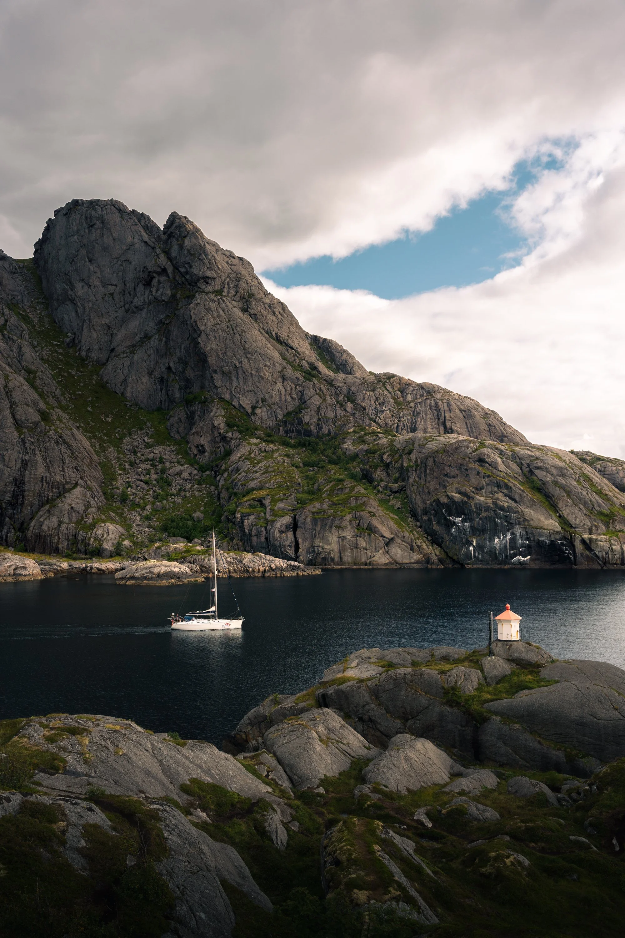 A scenic coastal landscape featuring a large rocky mountain, a calm body of water, a white sailboat, and a small lighthouse at the shoreline with a cloudy sky and a patch of blue sky peeking through.