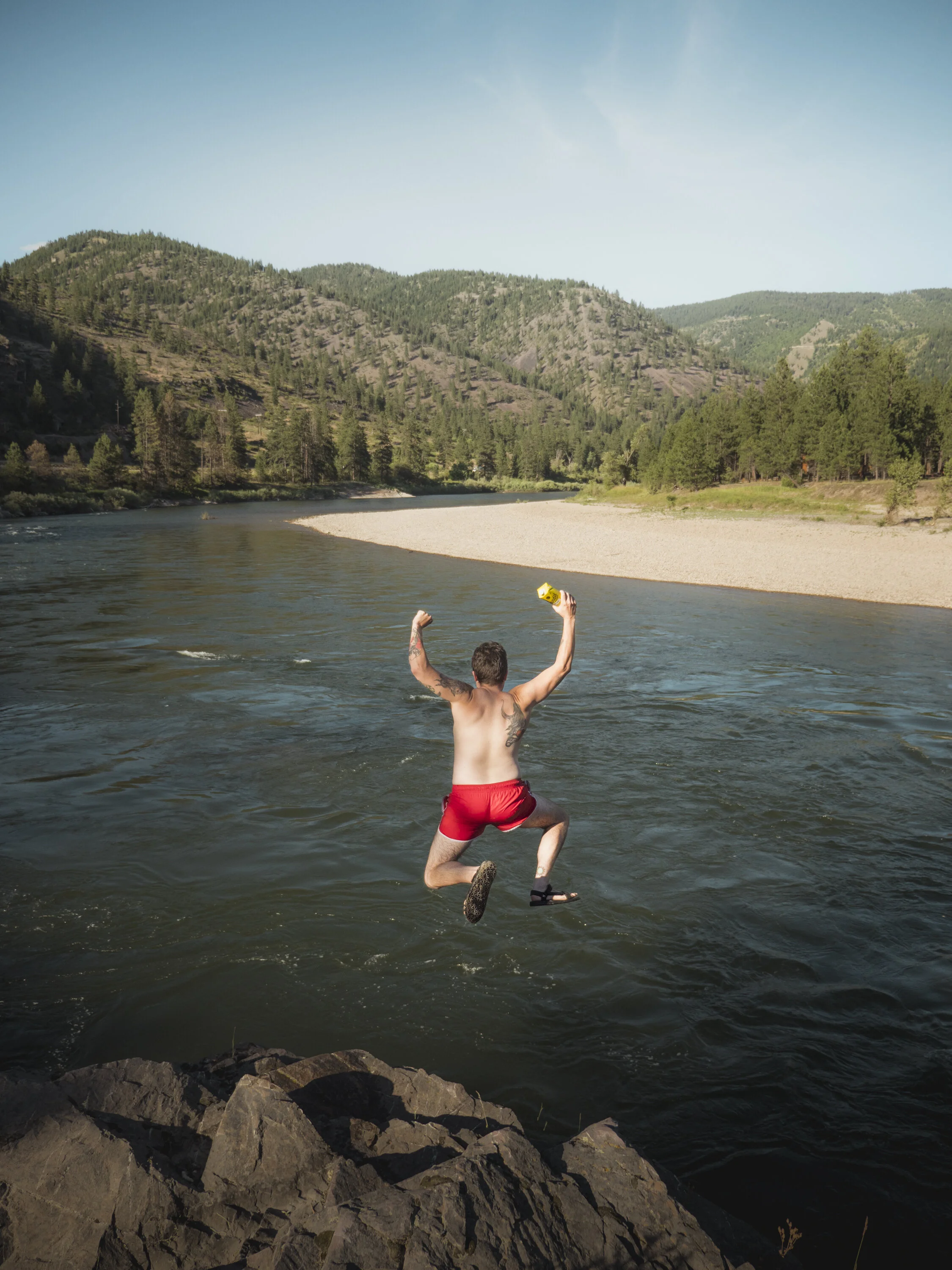 A person with tattoos and red shorts jumping into a river, surrounded by rocks and a forested landscape with hills and trees under a clear sky.