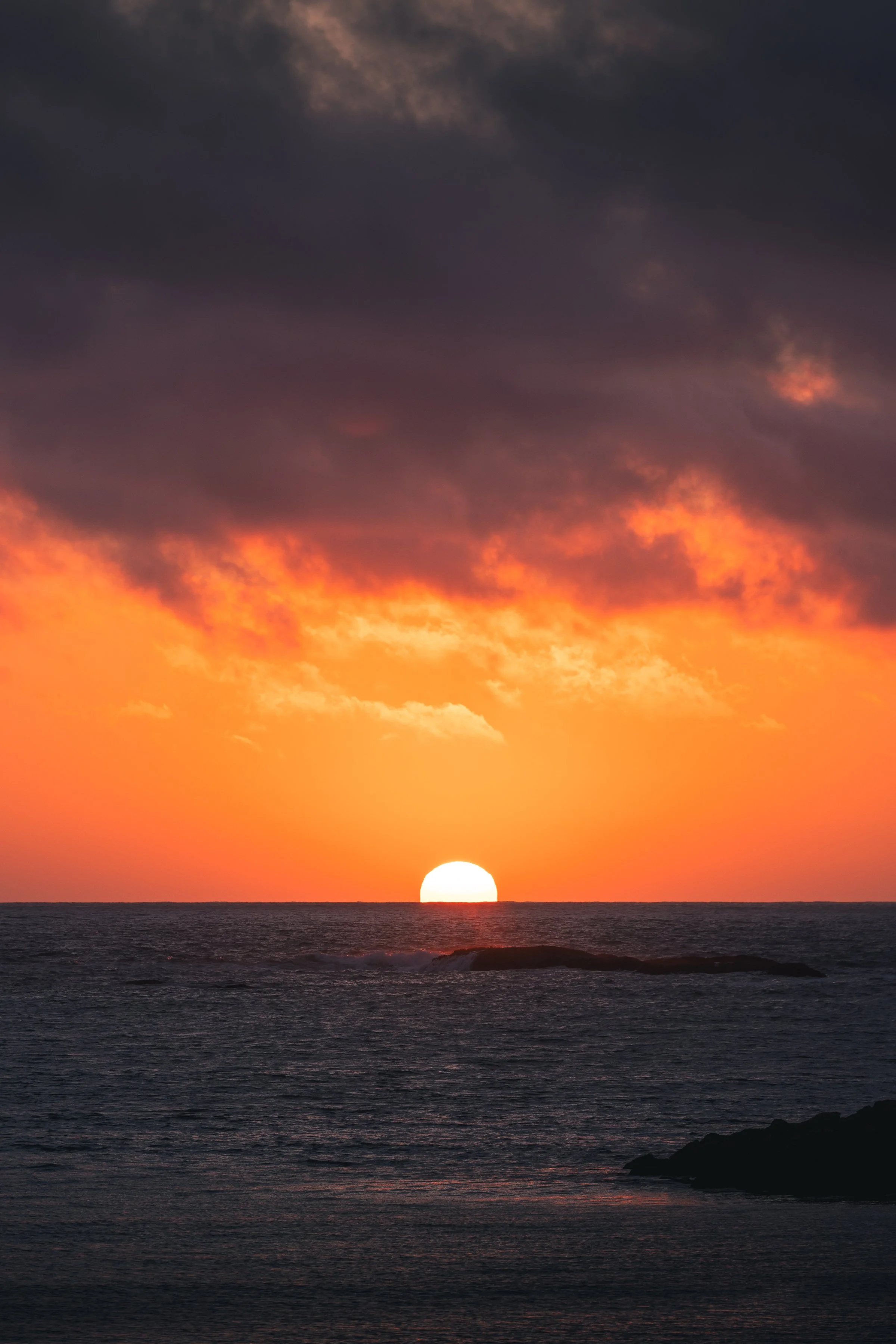 Sun setting over the ocean, with dark clouds in the sky and a silhouette of rocky shoreline in the foreground.