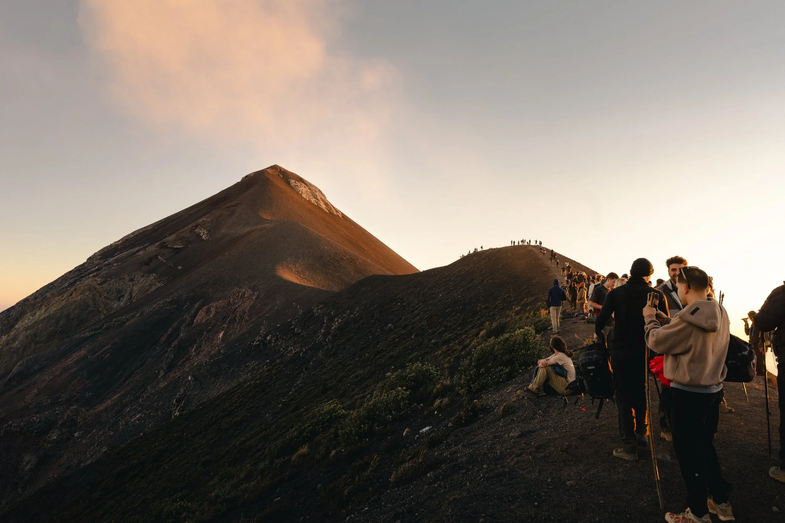Group of people hiking along a mountain trail during sunset or sunrise, with a mountain peak in the background.
