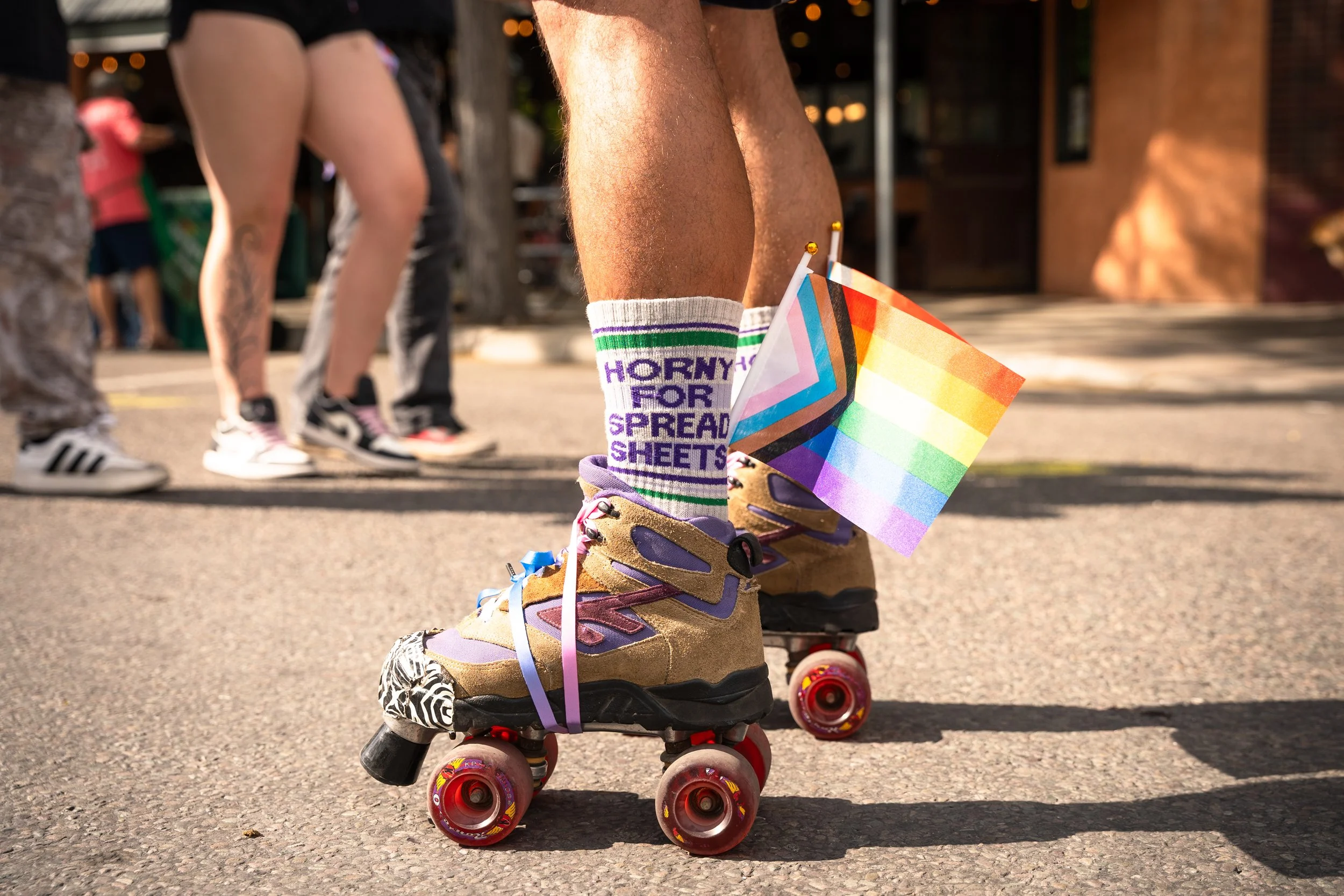 Close-up of a person wearing roller skates, purple and pink striped socks with the phrase 'HORNY FOR SPREAD SHEETS', and holding a rainbow pride flag. The background shows other skaters and people in an outdoor setting.