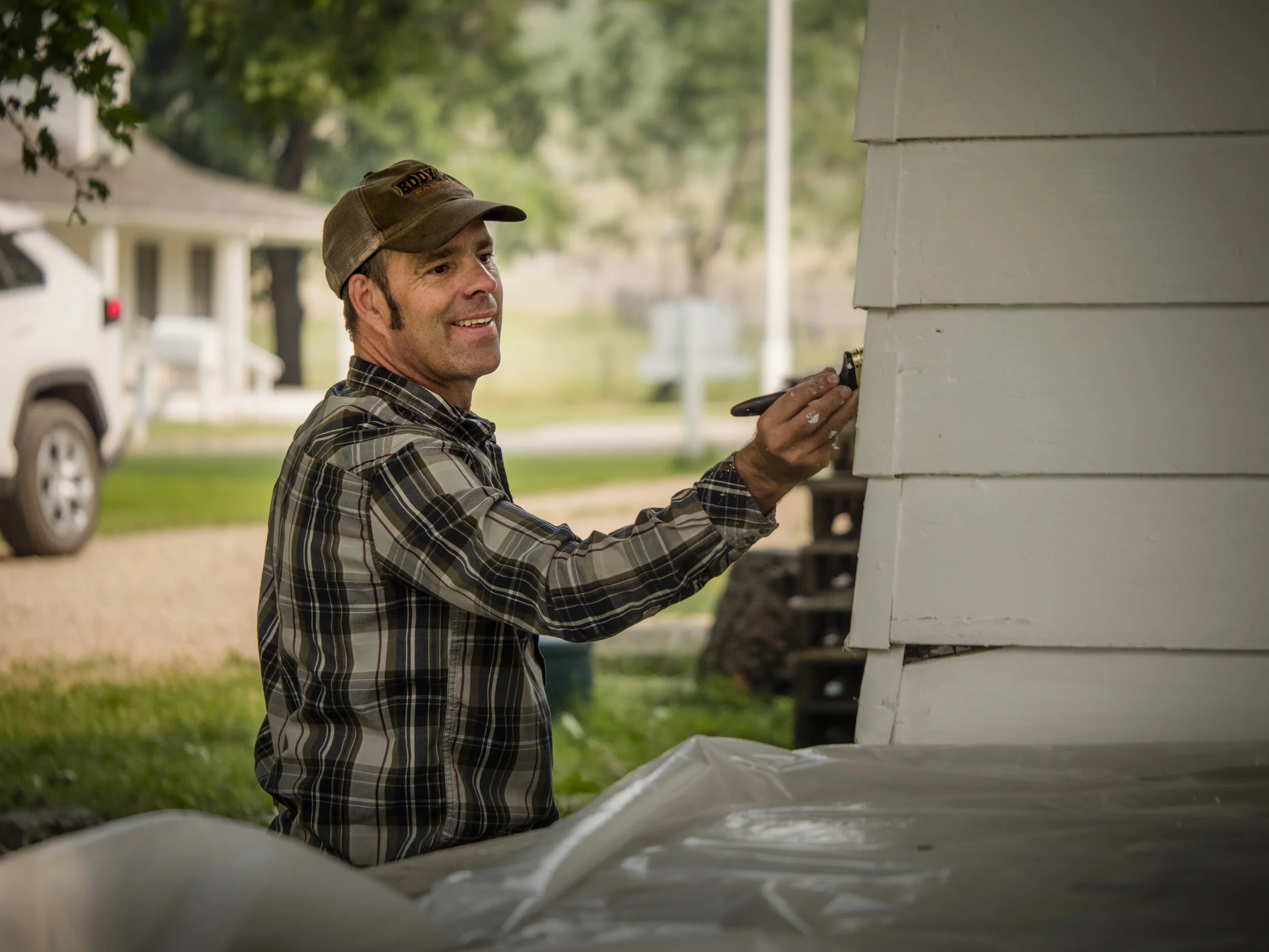 A man wearing a plaid shirt and a baseball cap working on a white wooden structure outdoors, with trees and houses in the background.