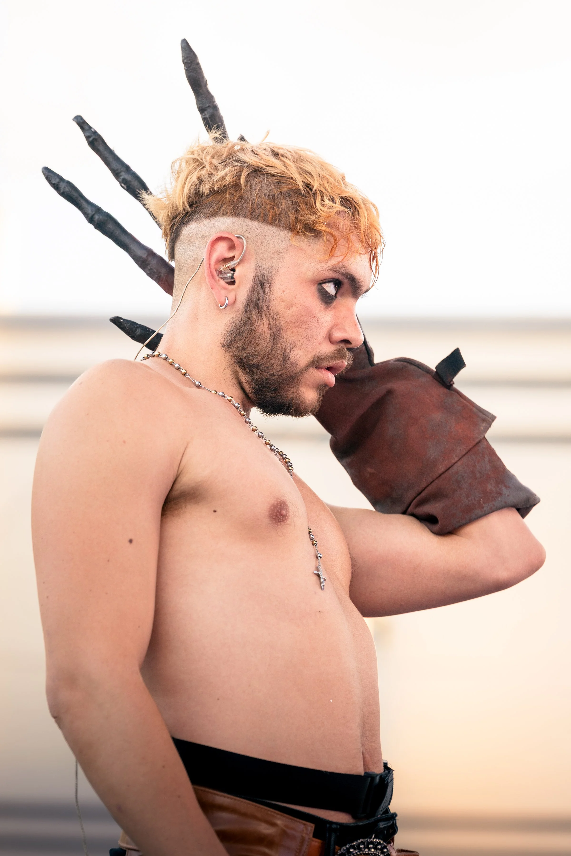 A shirtless young man with light skin, short wavy orange hair, beard, and multiple earrings, wearing a chain necklace and black pants with a belt, is carrying a large, rusty metal pipe on his shoulder. The background is blurred with neutral tones.