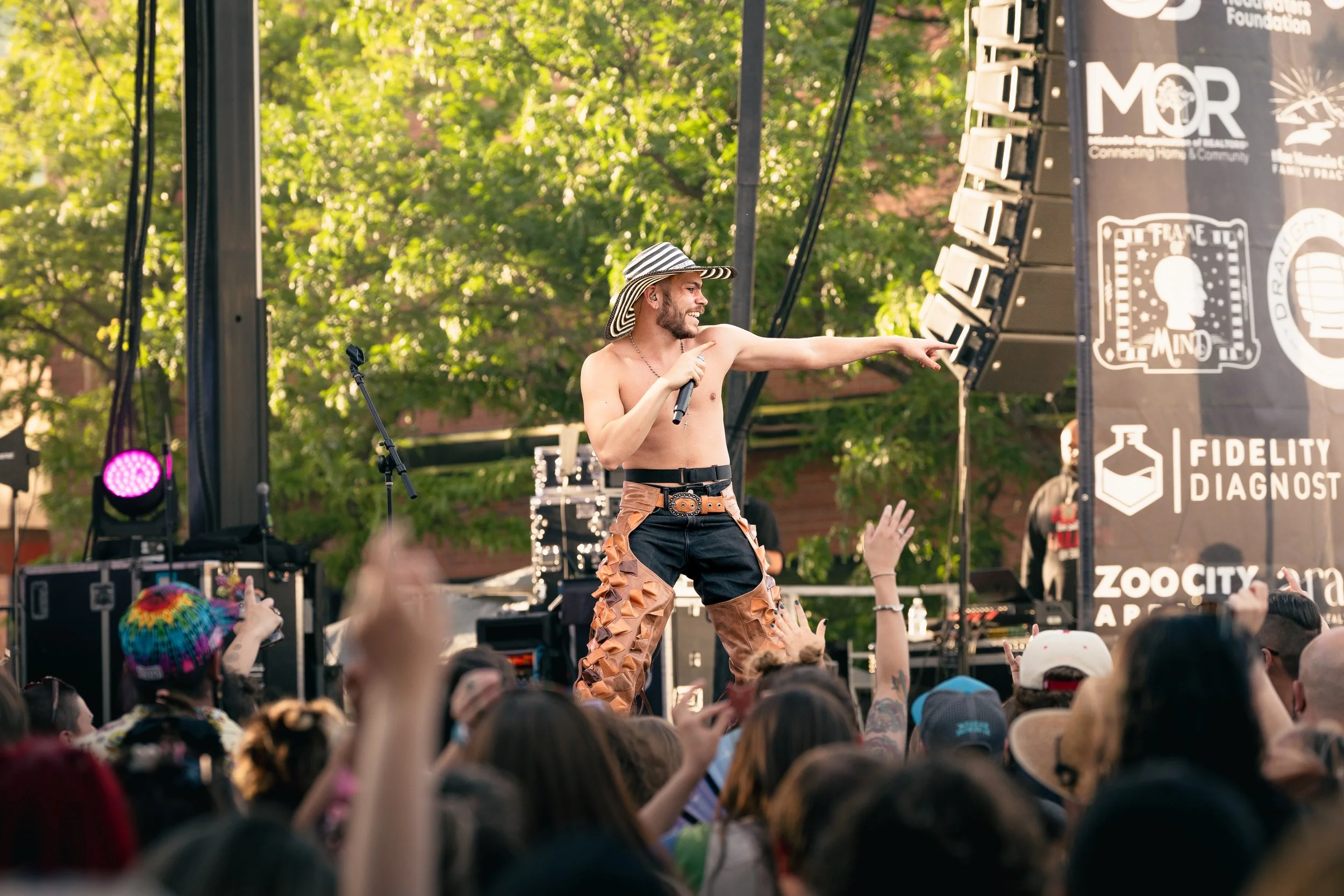 A shirtless male performer wearing a large striped sun hat, sunglasses, and decorated pants performing on an outdoor stage with a microphone in hand. The audience is visible with raised hands, some wearing hats and colorful accessories. The stage has