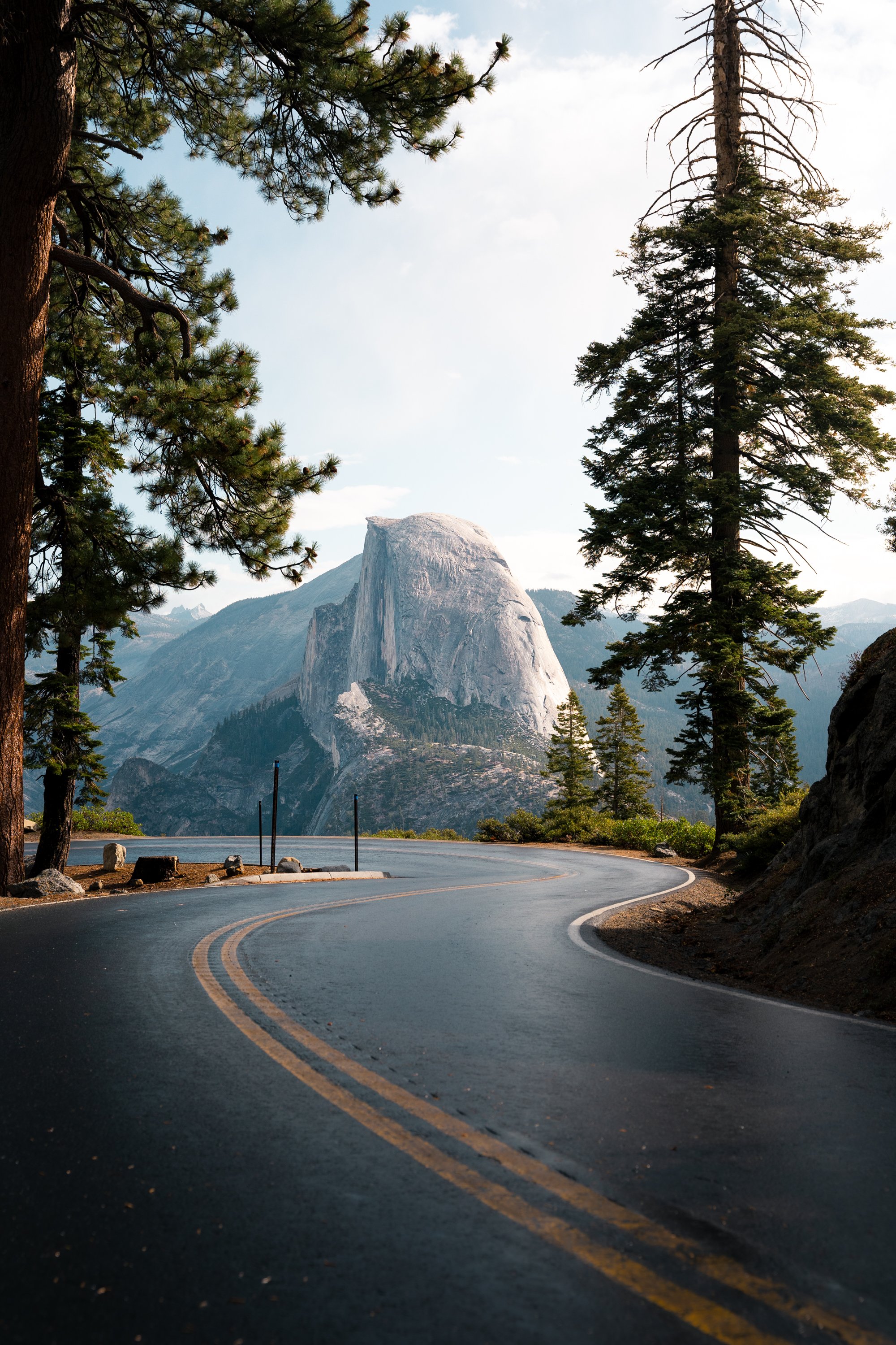 A winding mountain road with double yellow lines, bordered by pine trees on the left and right, with a large granite monolith mountain in the background under a partly cloudy sky.