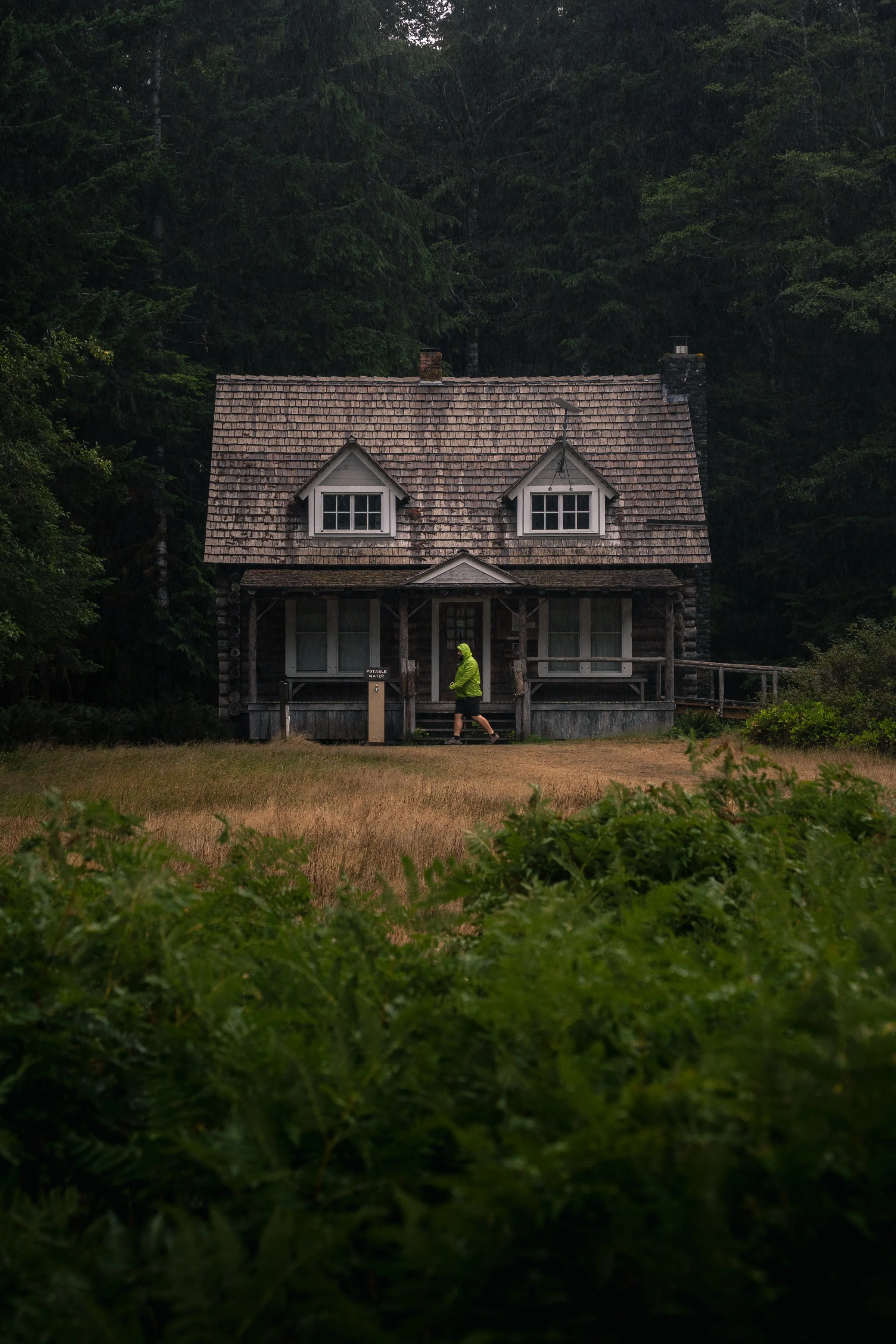 A rustic house with a shingled roof and dormer windows is situated in a forested area. A person in a bright green jacket is walking in front of the house. Lush greenery surrounds the scene.