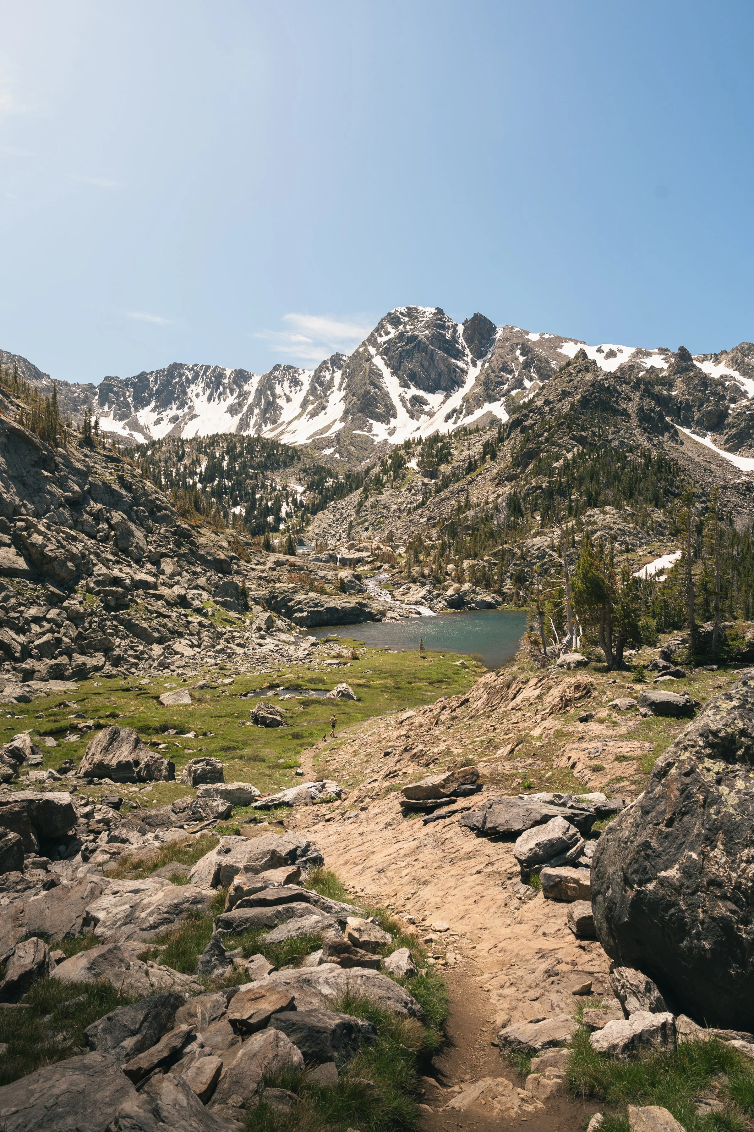 A hiking trail leading through a mountainous landscape with snow-capped peaks, a small lake, green vegetation, and a clear blue sky.