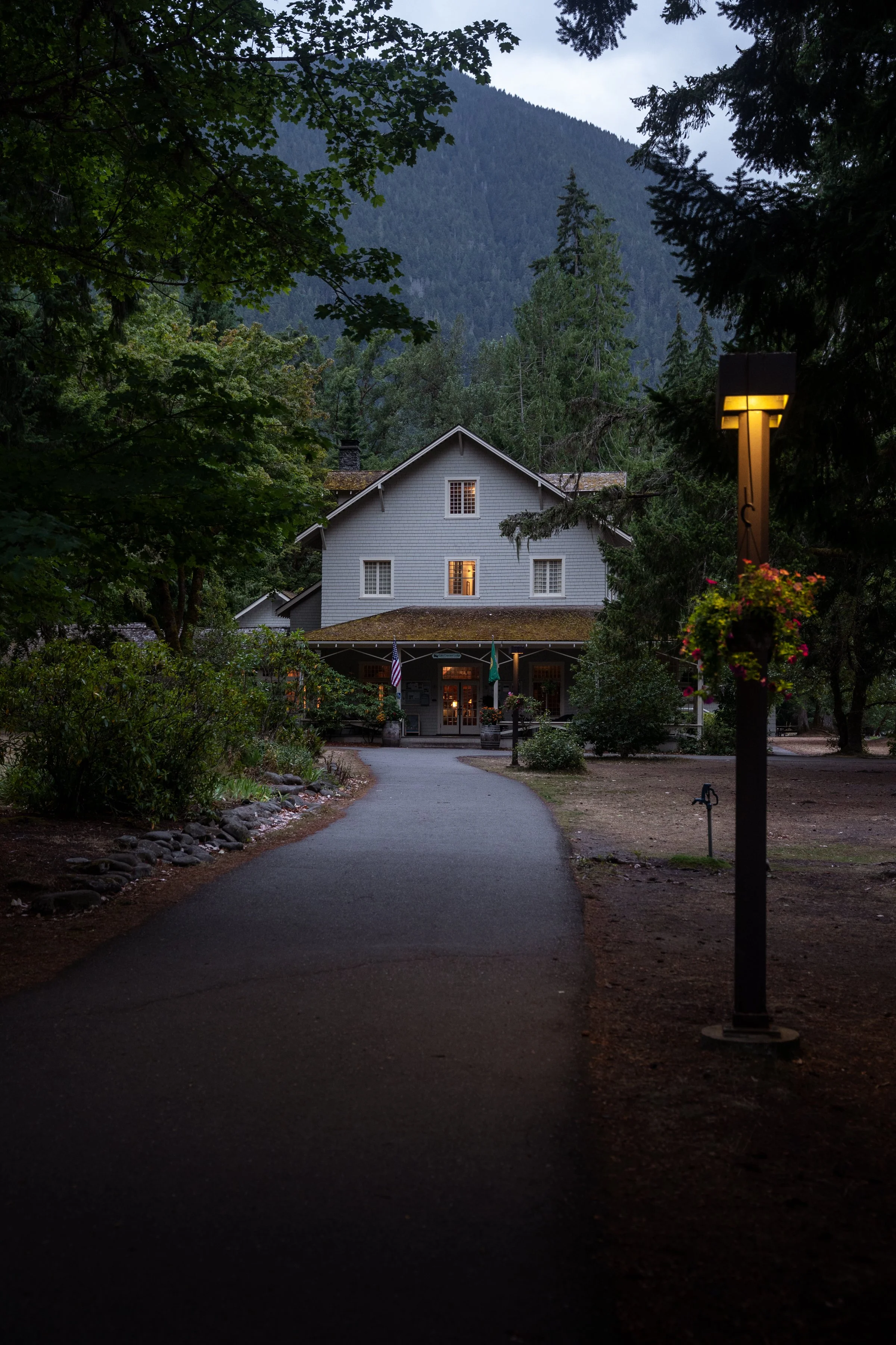 A cozy gray house with a porch, surrounded by trees and mountains in the background, during dusk with a streetlamp lit nearby.
