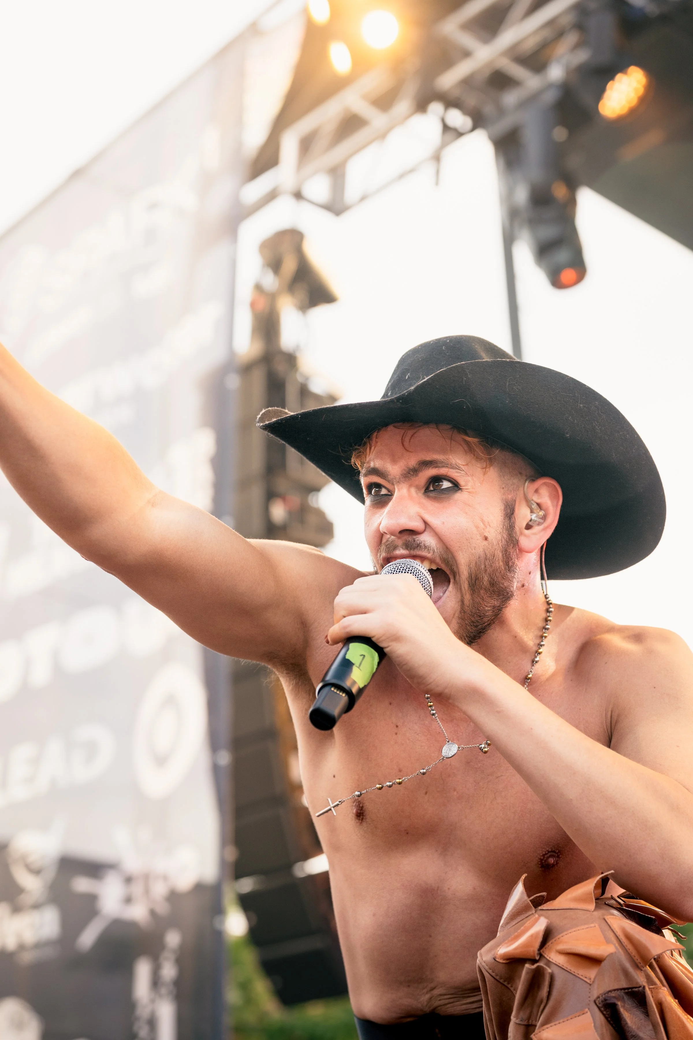 A shirtless man with a black cowboy hat singing into a microphone on an outdoor stage, holding up his right arm, with stage lights and a large screen in the background.