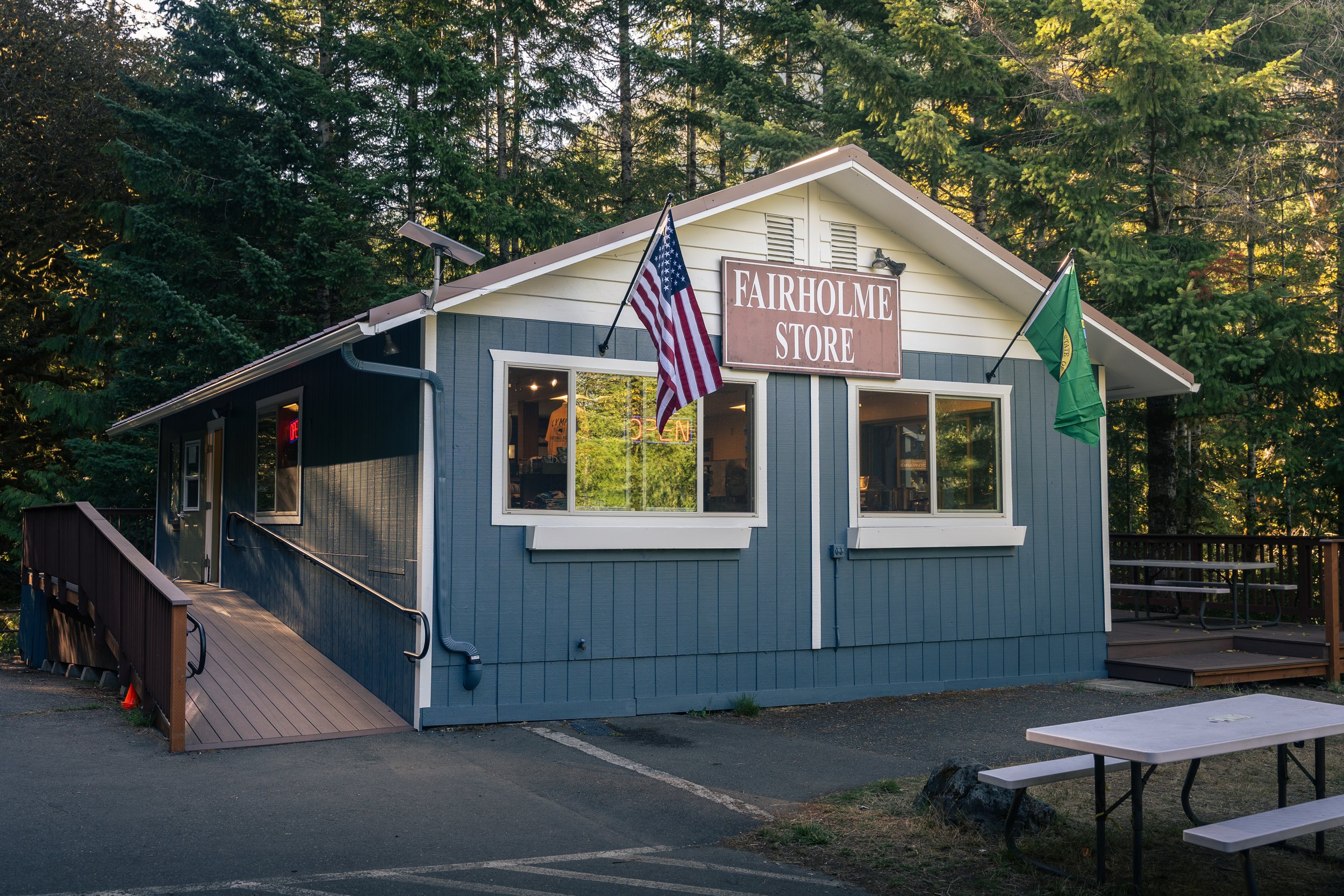 Small blue building labeled 'Fairholme Store' surrounded by tall pine trees, with American flag and green flag on poles outside, and a ramp leading to the entrance, indicating it is an accessible store in a wooded area.