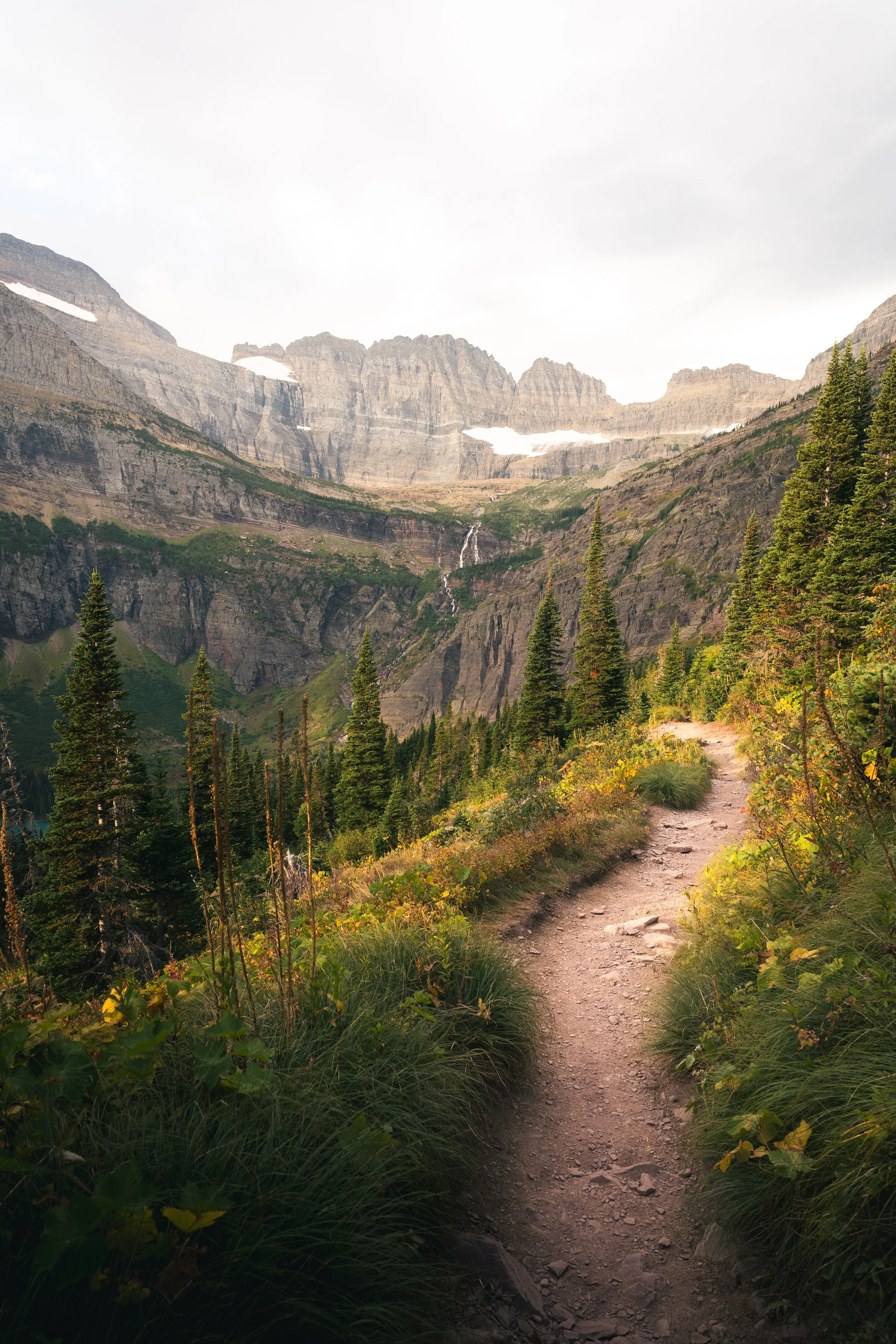 A rugged mountain trail winding through a valley with dense green pine trees, steep rocky mountains in the background, patches of snow, and a partly cloudy sky.