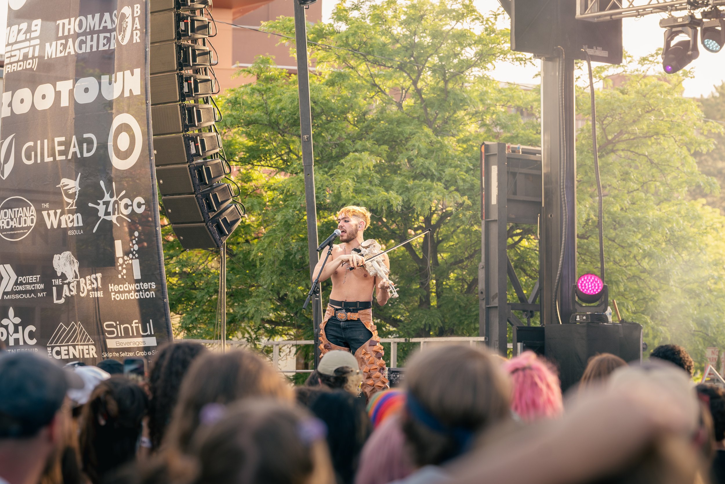 A shirtless performer with yellow hair and makeup, wearing patterned pants and a belt, holding a violin and speaking into a microphone on an outdoor stage during a concert, with a crowd in front, surrounded by trees and stage equipment.