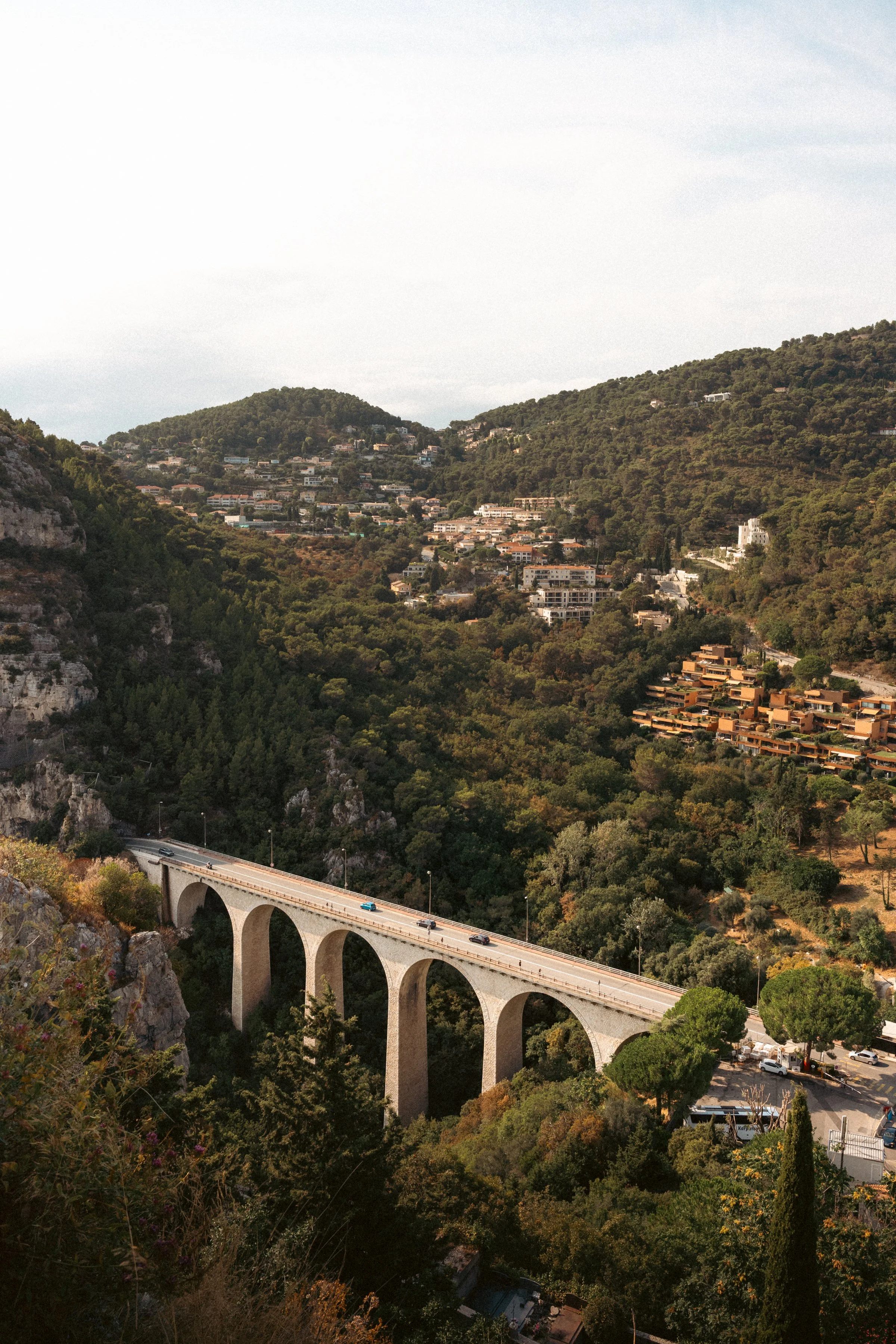 Scenic view of a mountainous area with many residential houses on green hillsides. A large stone arched bridge crosses a valley in the foreground, with a few cars traveling on it. Dense trees cover much of the landscape, and the sky is partly cloudy.