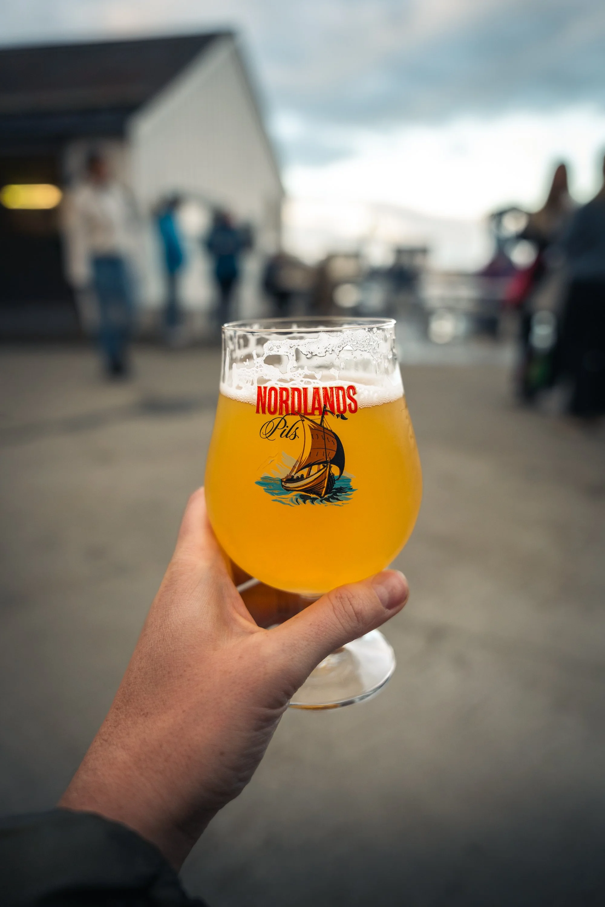 A person holding a glass of beer with a logo 'Nordlands Pils' featuring a sailing ship, in an outdoor setting with a blurred background of people and a building.