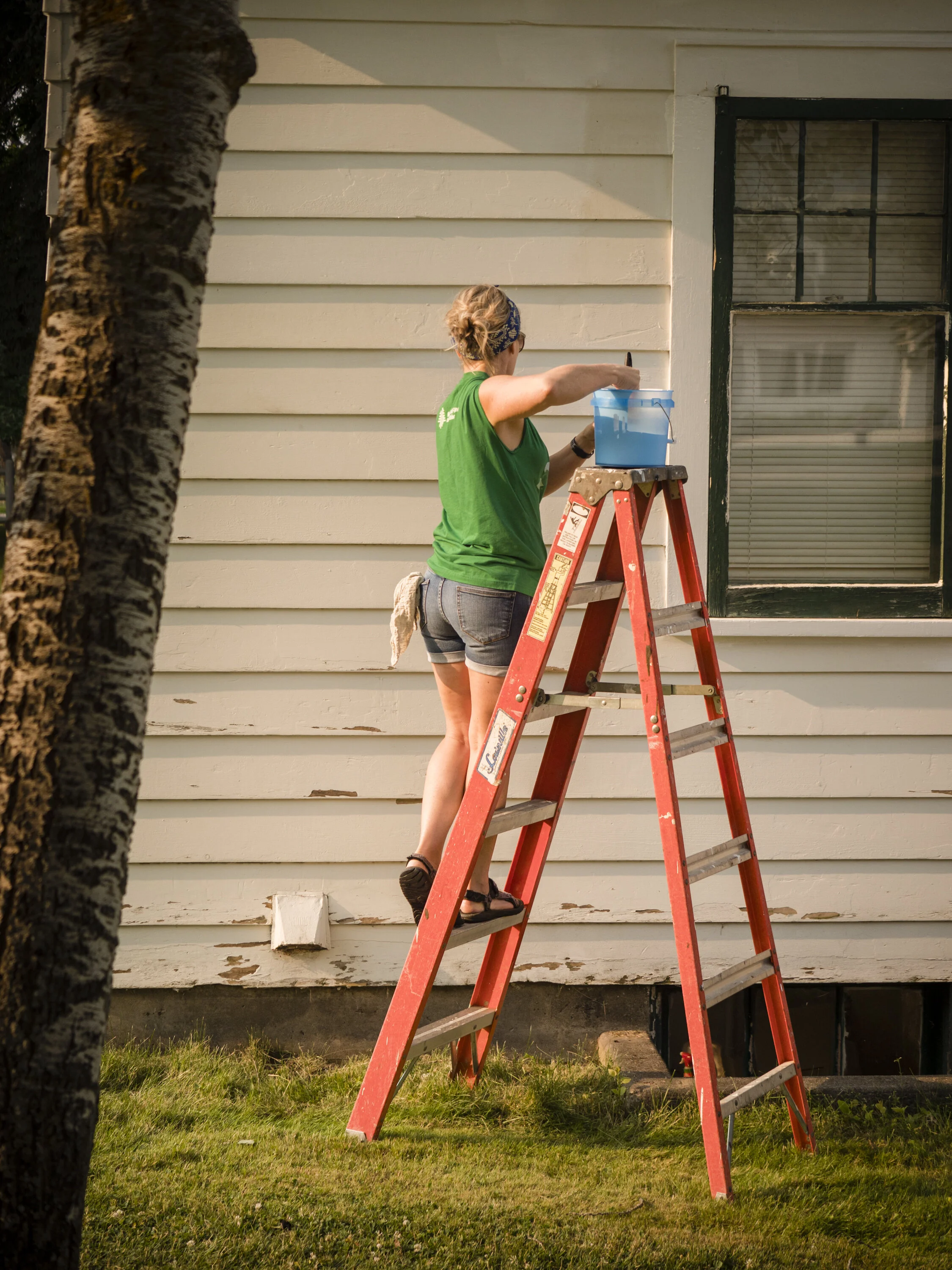 Woman standing on a red ladder painting the exterior wall of a house.