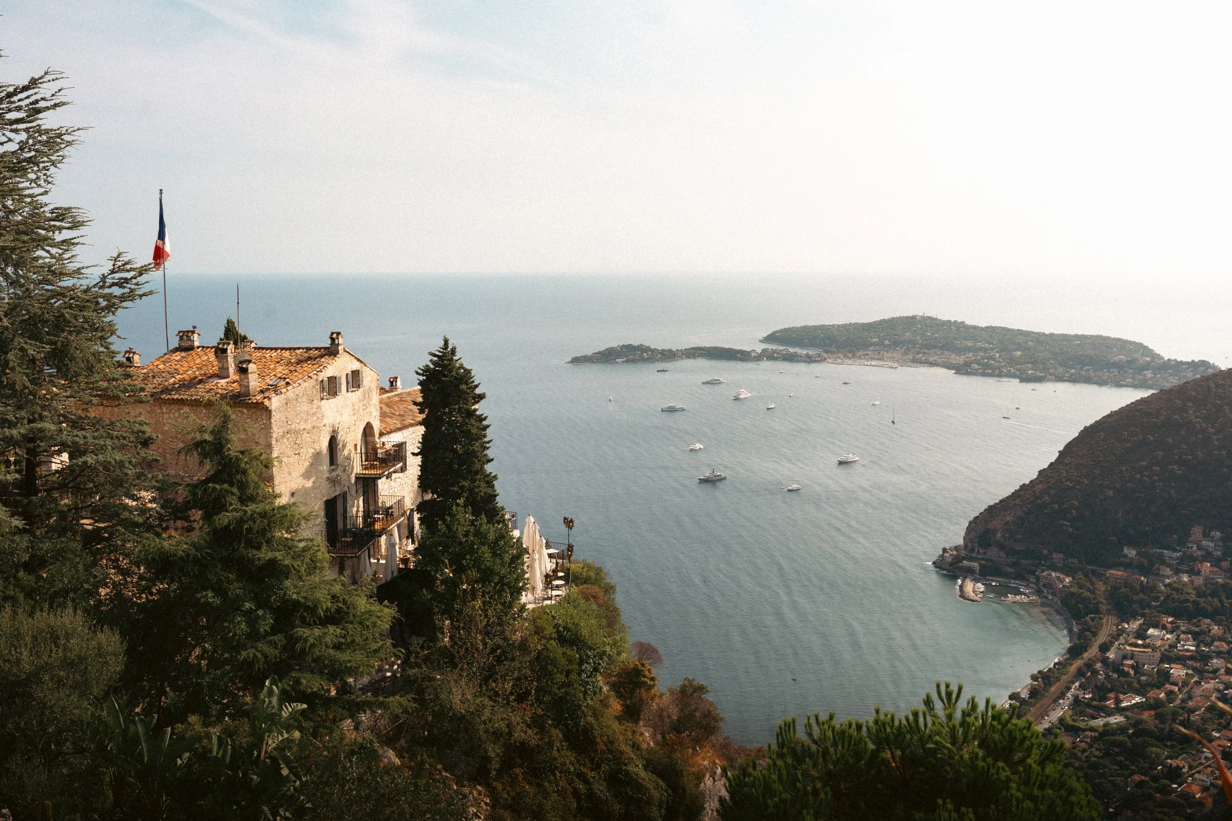 A scenic coastal view with historic buildings, trees, and a harbor with boats, overlooking the ocean and distant islands.