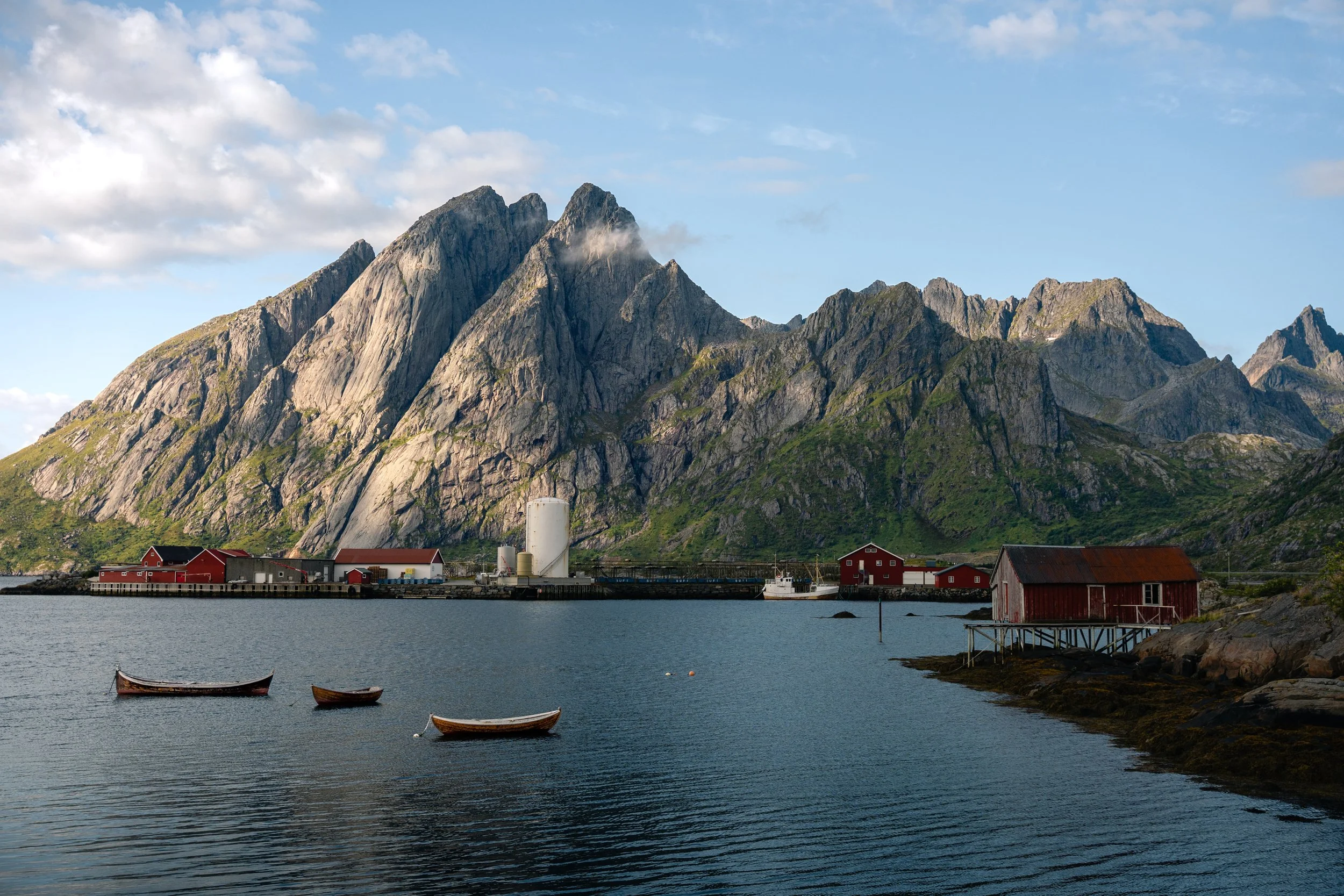 A coastal town with red and gray buildings along the water's edge, two small wooden boats floating on the water, and large rocky mountains in the background under a partly cloudy sky.
