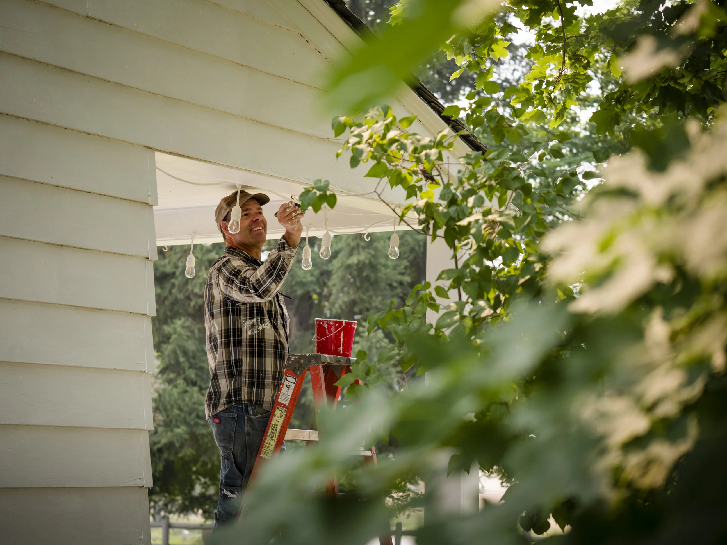 A man standing on a ladder, hanging string lights on the porch of a house, surrounded by green leaves.