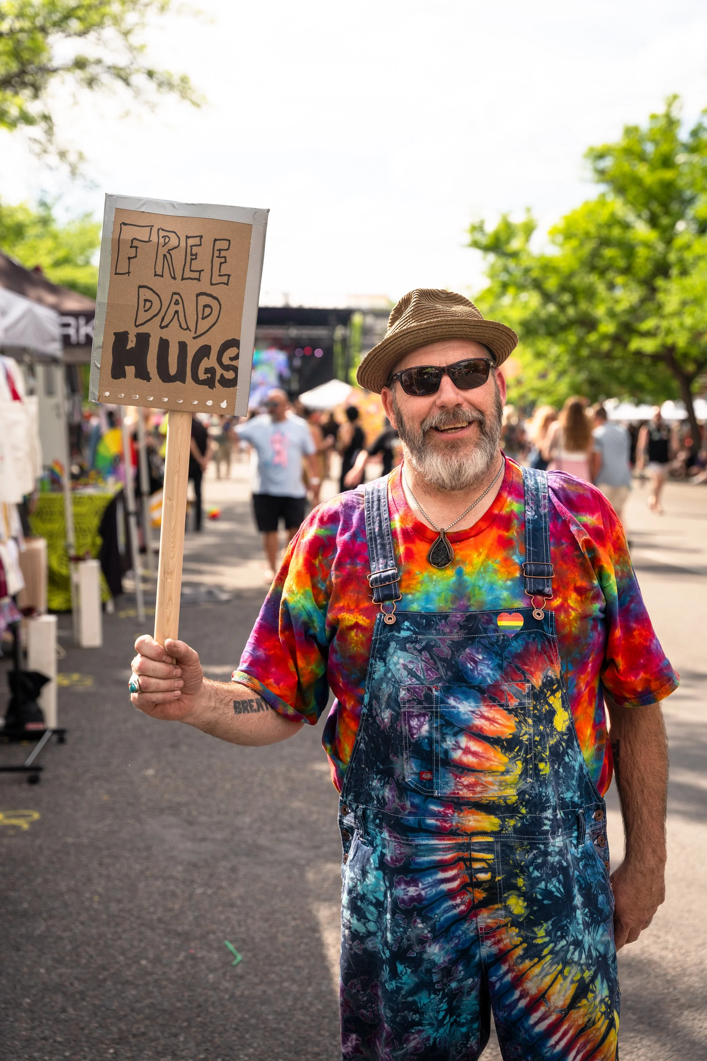 A man with a gray beard wearing a colorful tie-dye shirt, denim overalls, sunglasses, and a hat, holding a handmade sign that says 'Free Dad Hugs' at an outdoor event or festival with trees and people in the background