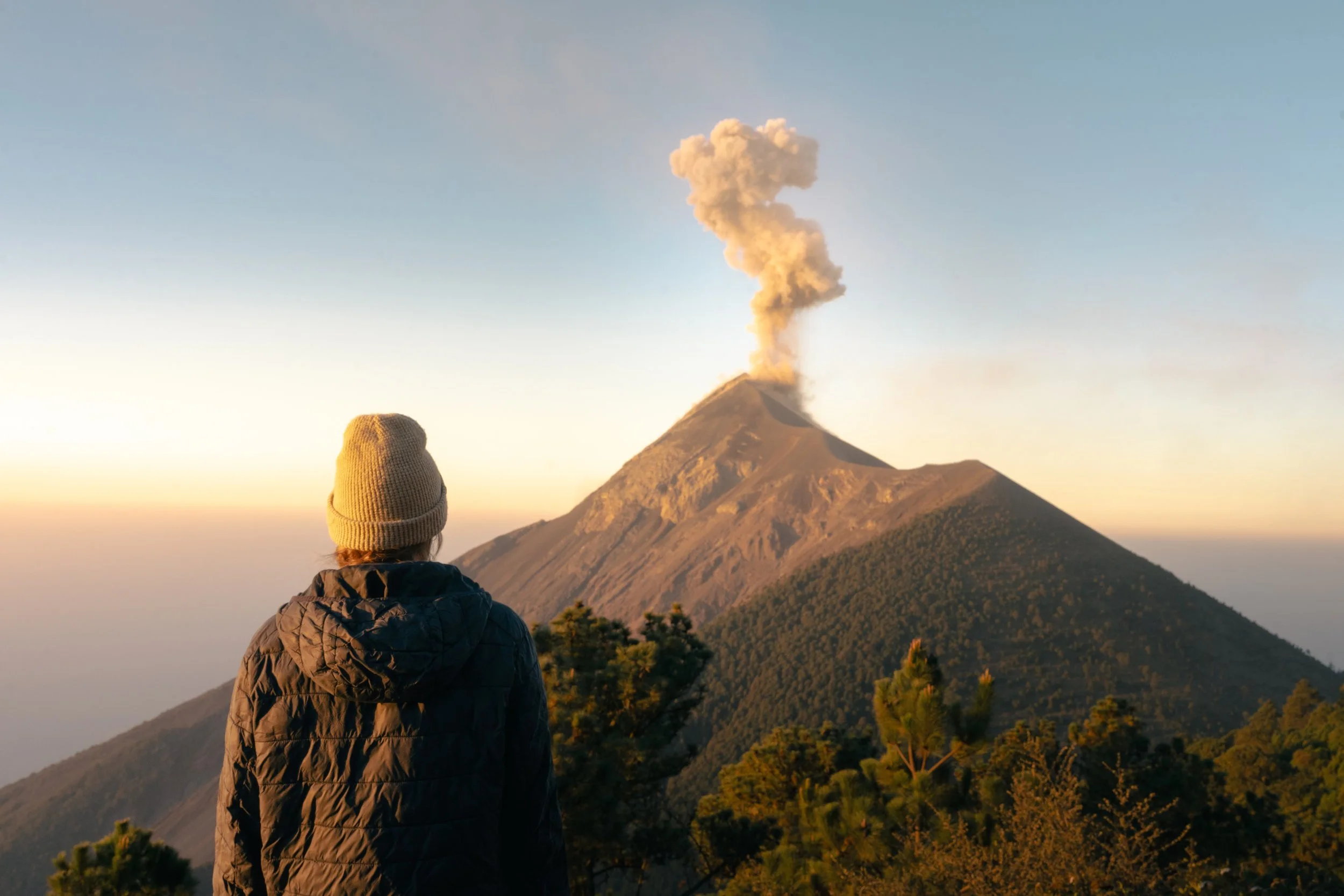 Person wearing a hat and jacket looking at a volcanic eruption with ash and smoke rising from the volcano's peak, in the late afternoon or early evening.