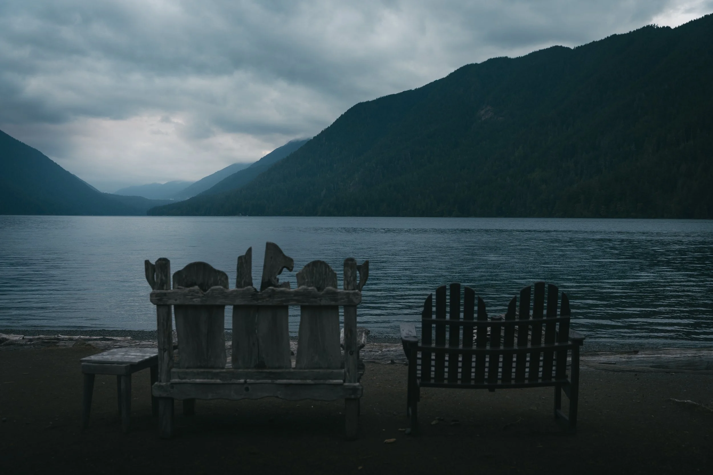 Three wooden chairs are set on a sandy shore overlooking a calm lake with mountains and cloudy sky in the background.