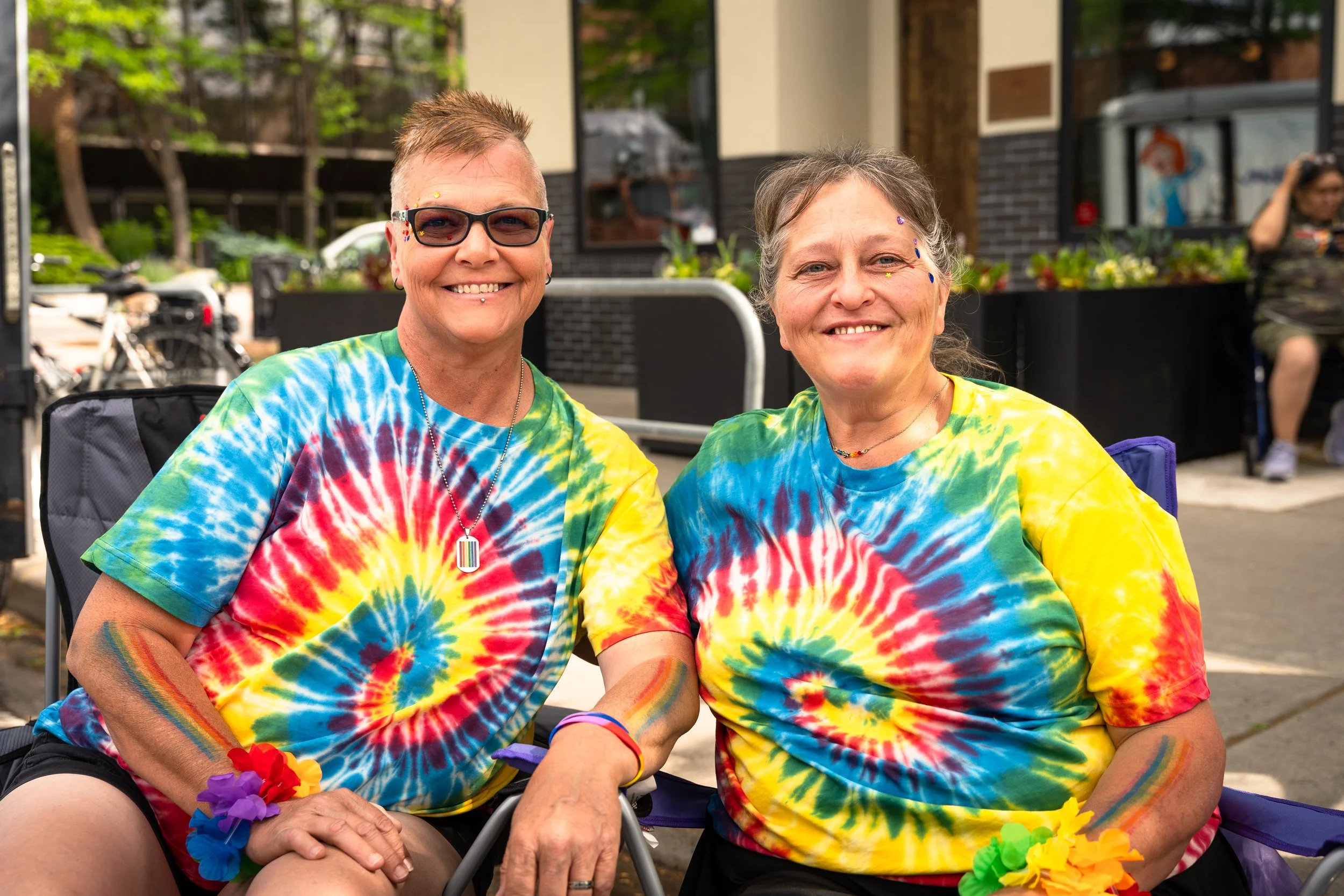 Two smiling women wearing rainbow tie-dye shirts and colorful floral accessories, sitting outdoors on a sunny day at a pride celebration, with buildings and other people in the background.