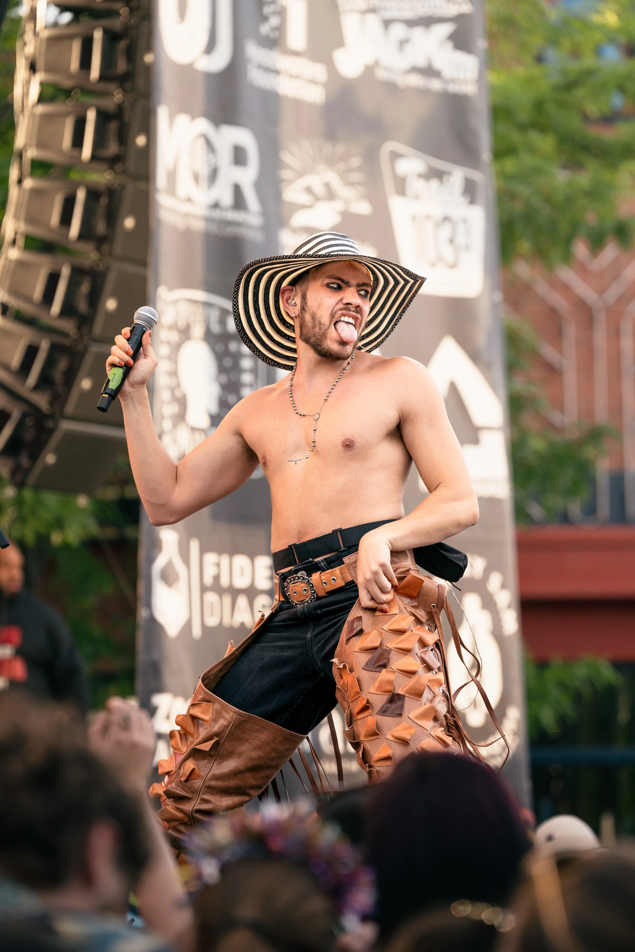 A shirtless man with a large striped sun hat and brown leather pants with laces, standing on stage holding a microphone, making a playful face with his tongue out, at an outdoor event with a crowd and a banner in the background.