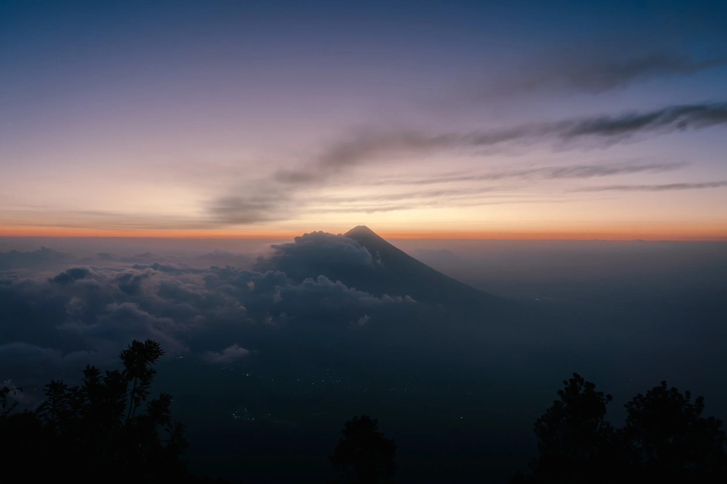 Sunset over a mountain with clouds, silhouetted trees in the foreground, and a transition of colors from orange to purple in the sky.