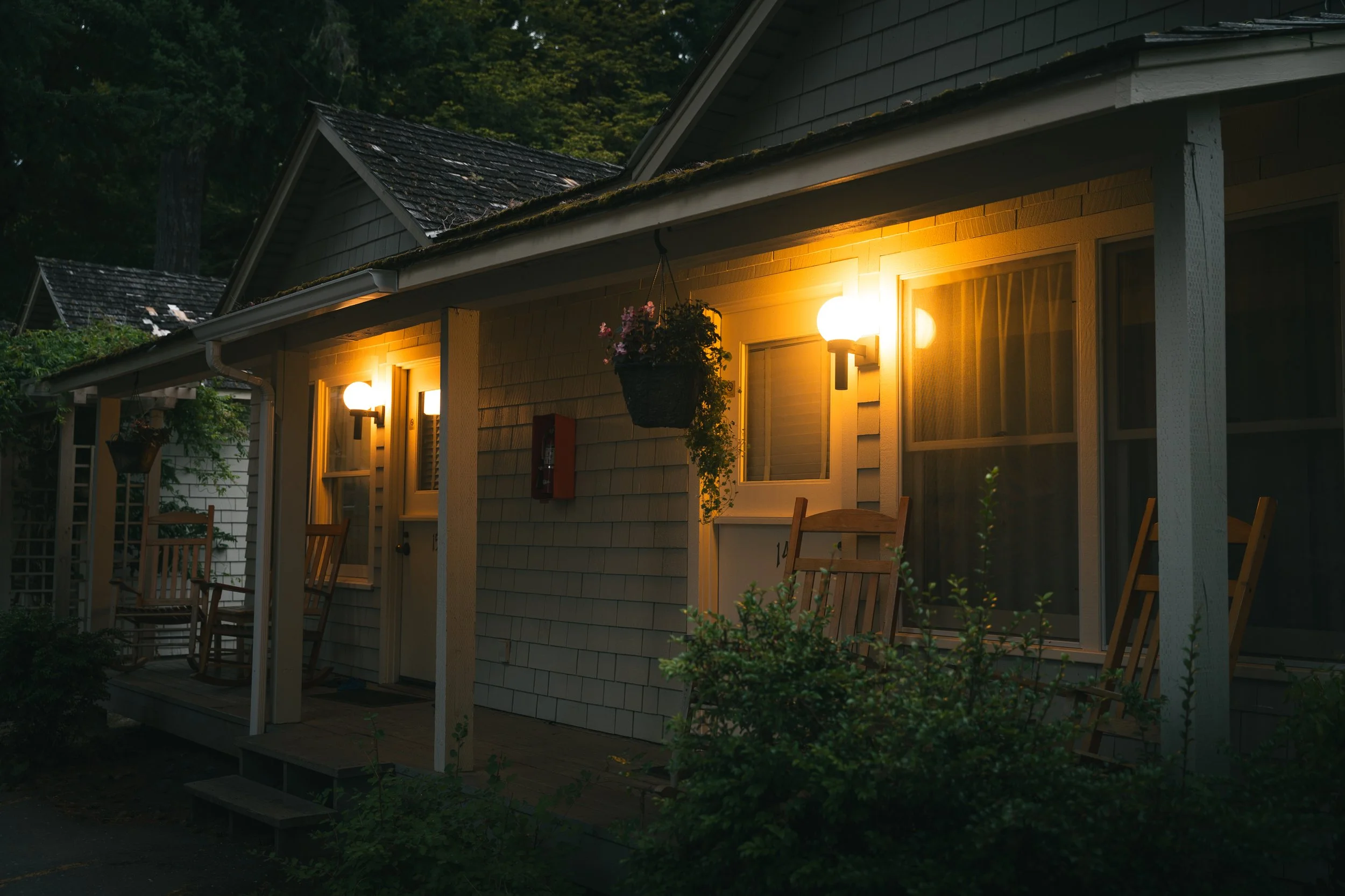 A house with a porch illuminated by exterior lights at dusk, surrounded by greenery and trees in the background.