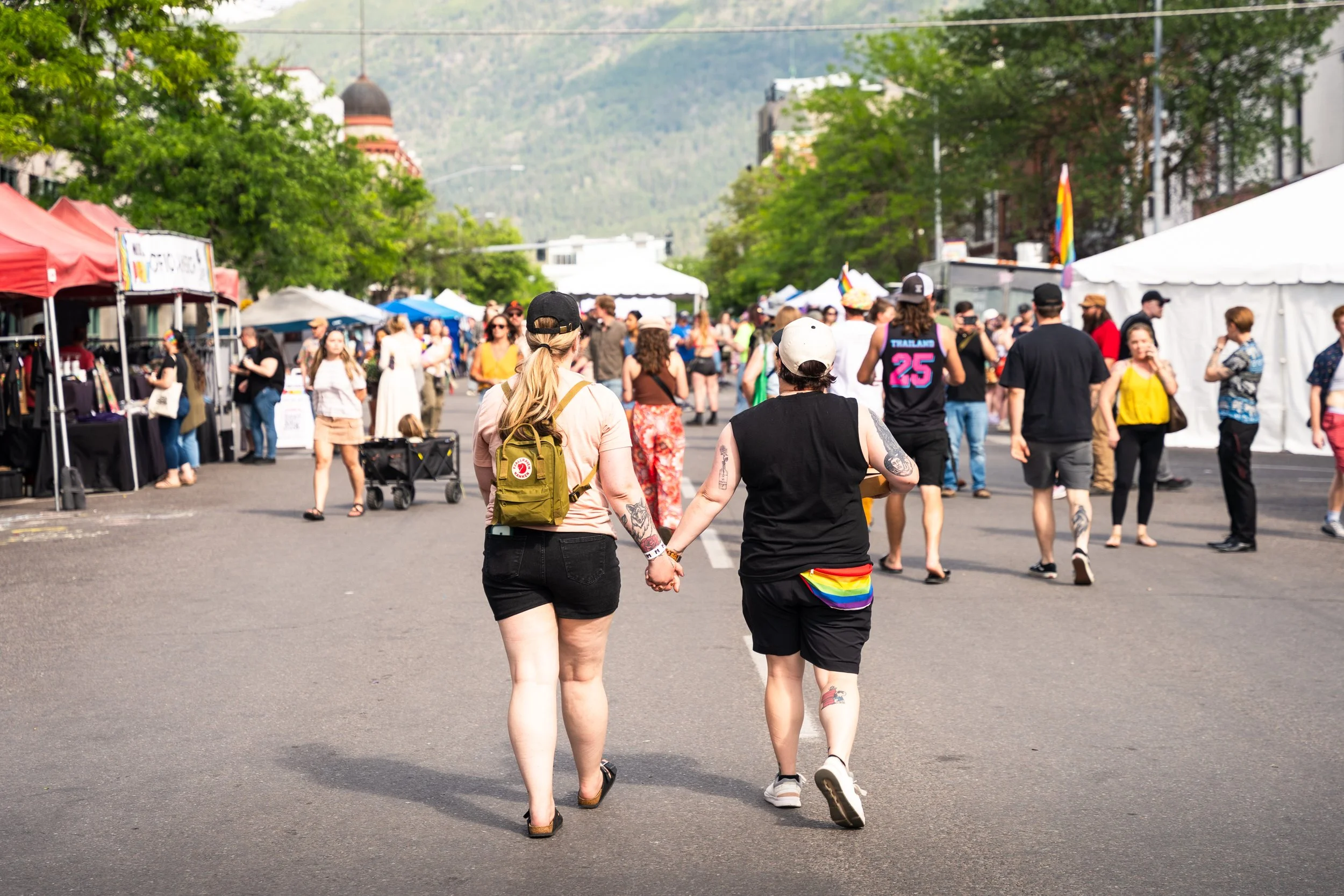 A crowd of people walking at an outdoor event with tents and trees, some wearing rainbow accessories and pride flags.