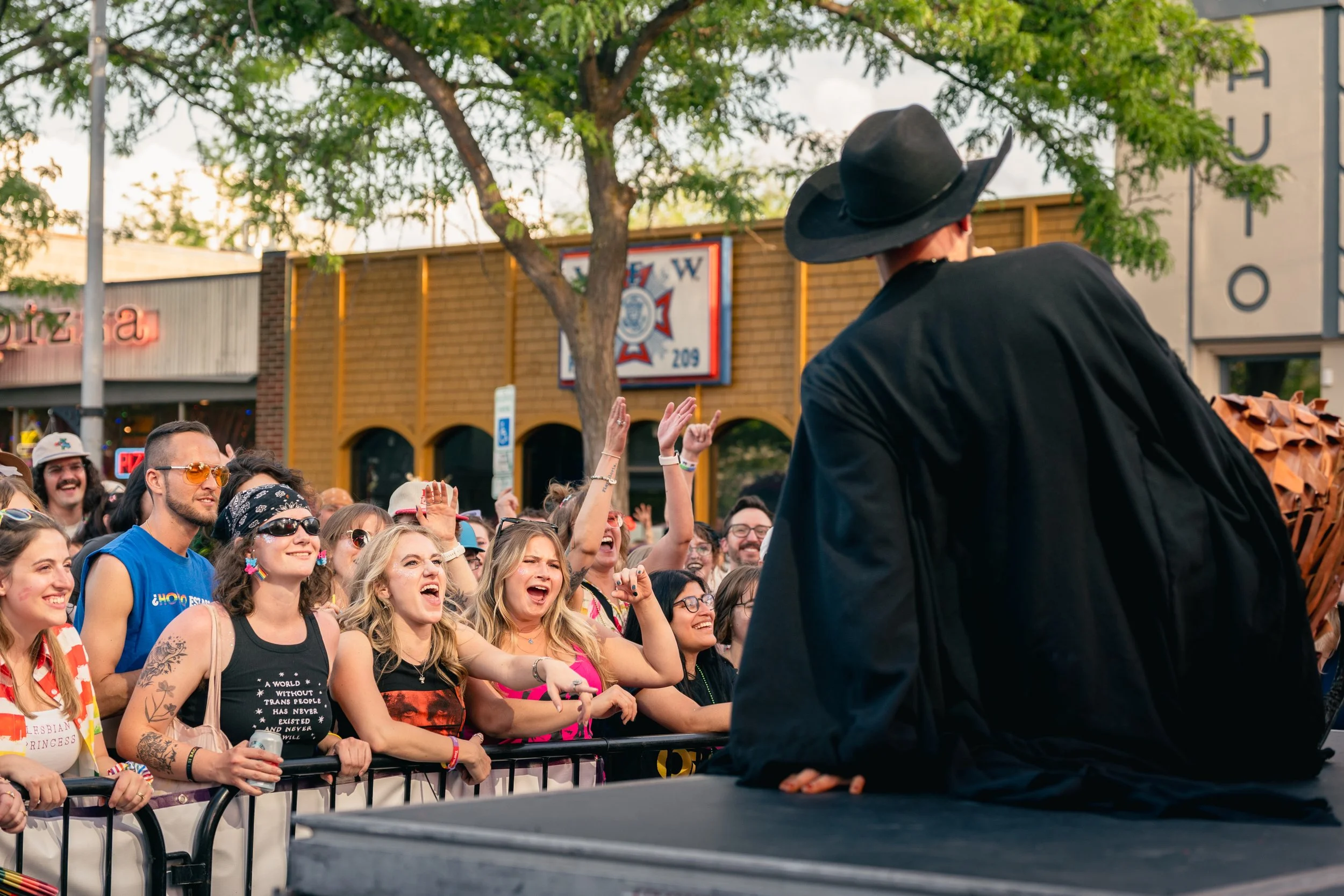 A crowd of people at an outdoor event, with many smiling and some raising their hands. A person wearing a black hat and black clothing is in the foreground, facing the crowd. In the background, there are trees and buildings.