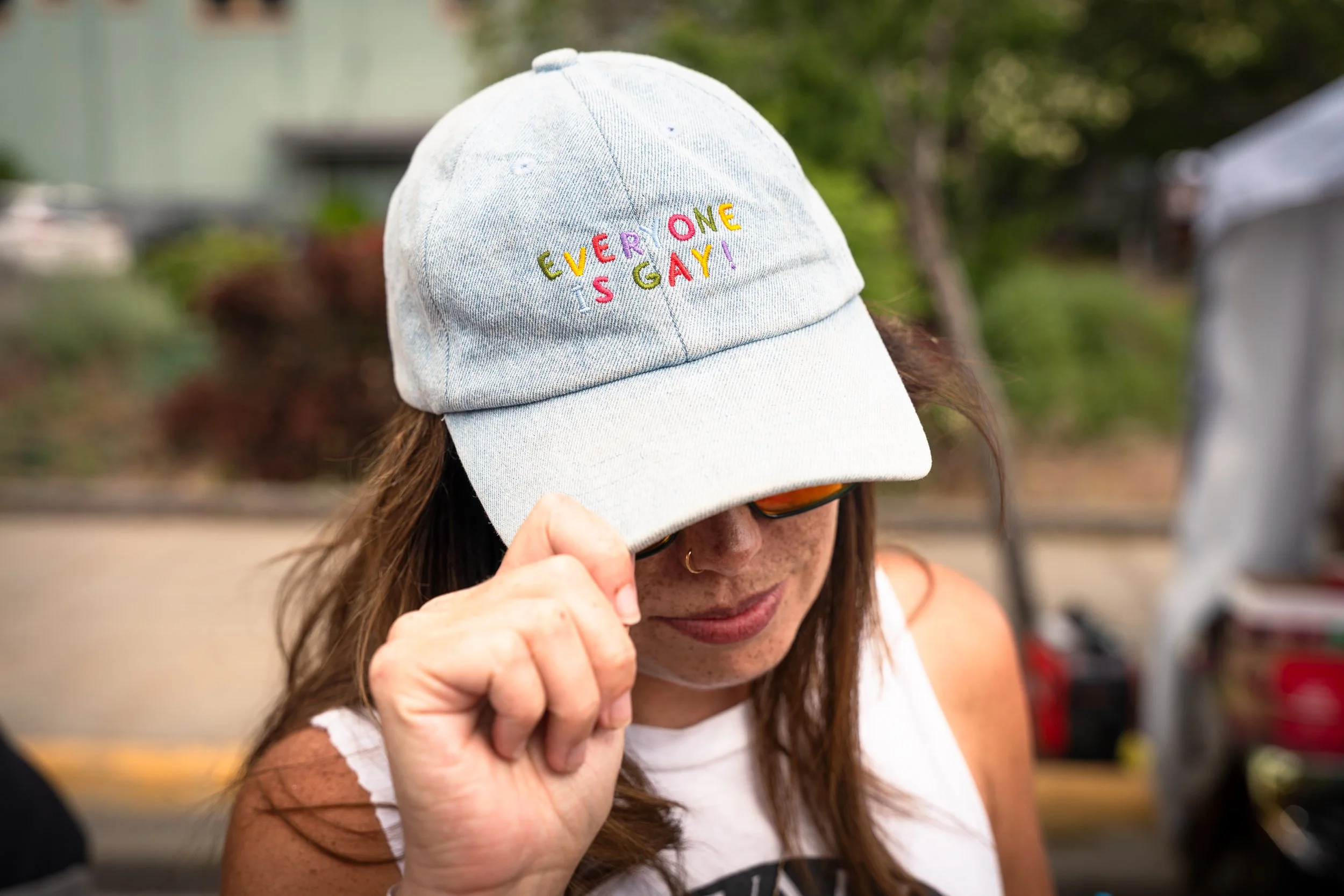 A woman wearing a grey baseball cap with the embroidered text 'Everyone is Gay' in rainbow colors, adjusting the cap with her hand.