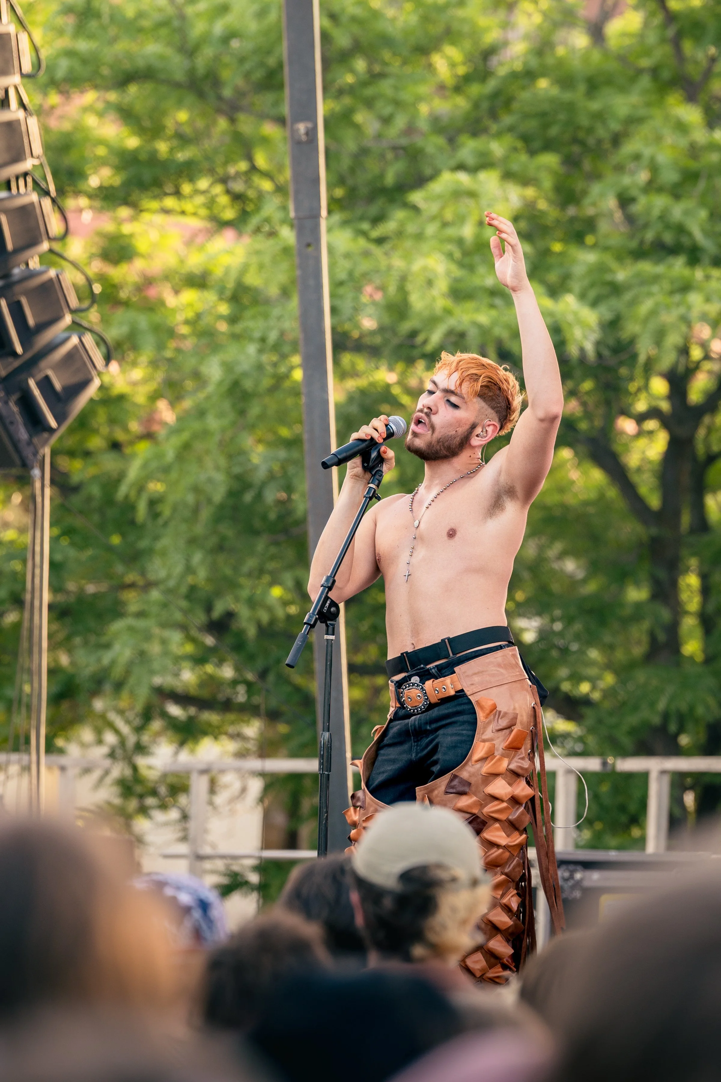 A shirtless male performer with orange hair singing into a microphone on an outdoor stage, raising his hand, with a crowd watching in front and green trees in the background.