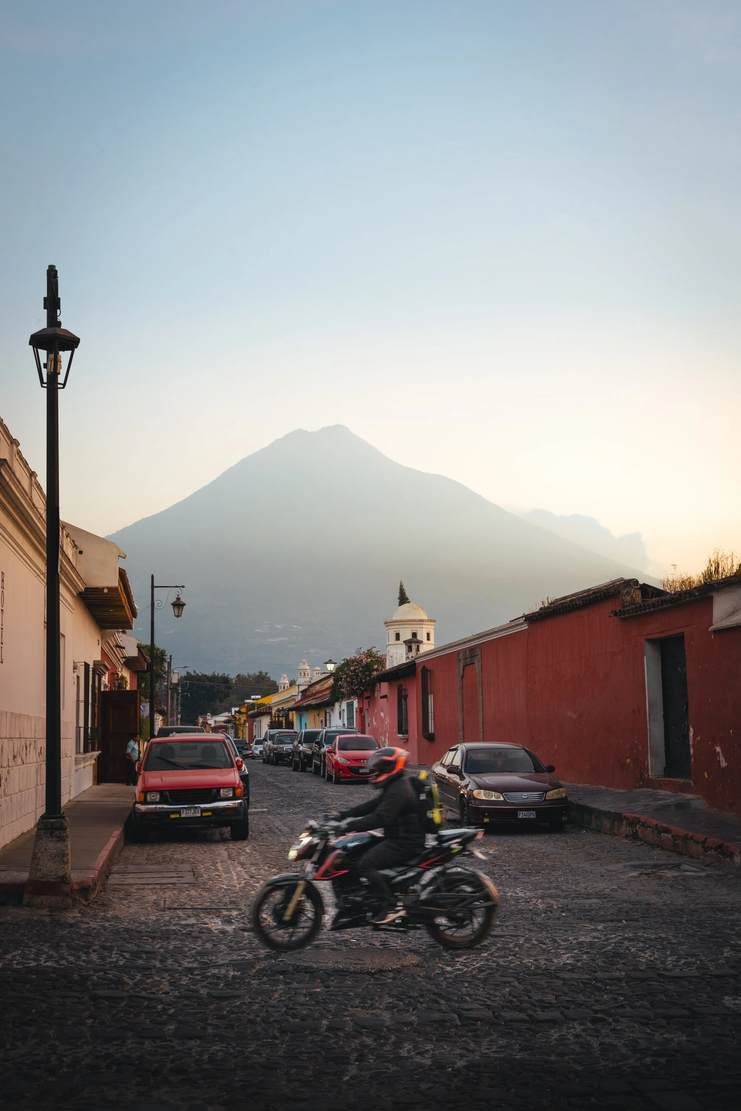 A cobblestone street in a colorful town with parked cars, a moving motorcycle, and colonial-style buildings under a mountain in the background.