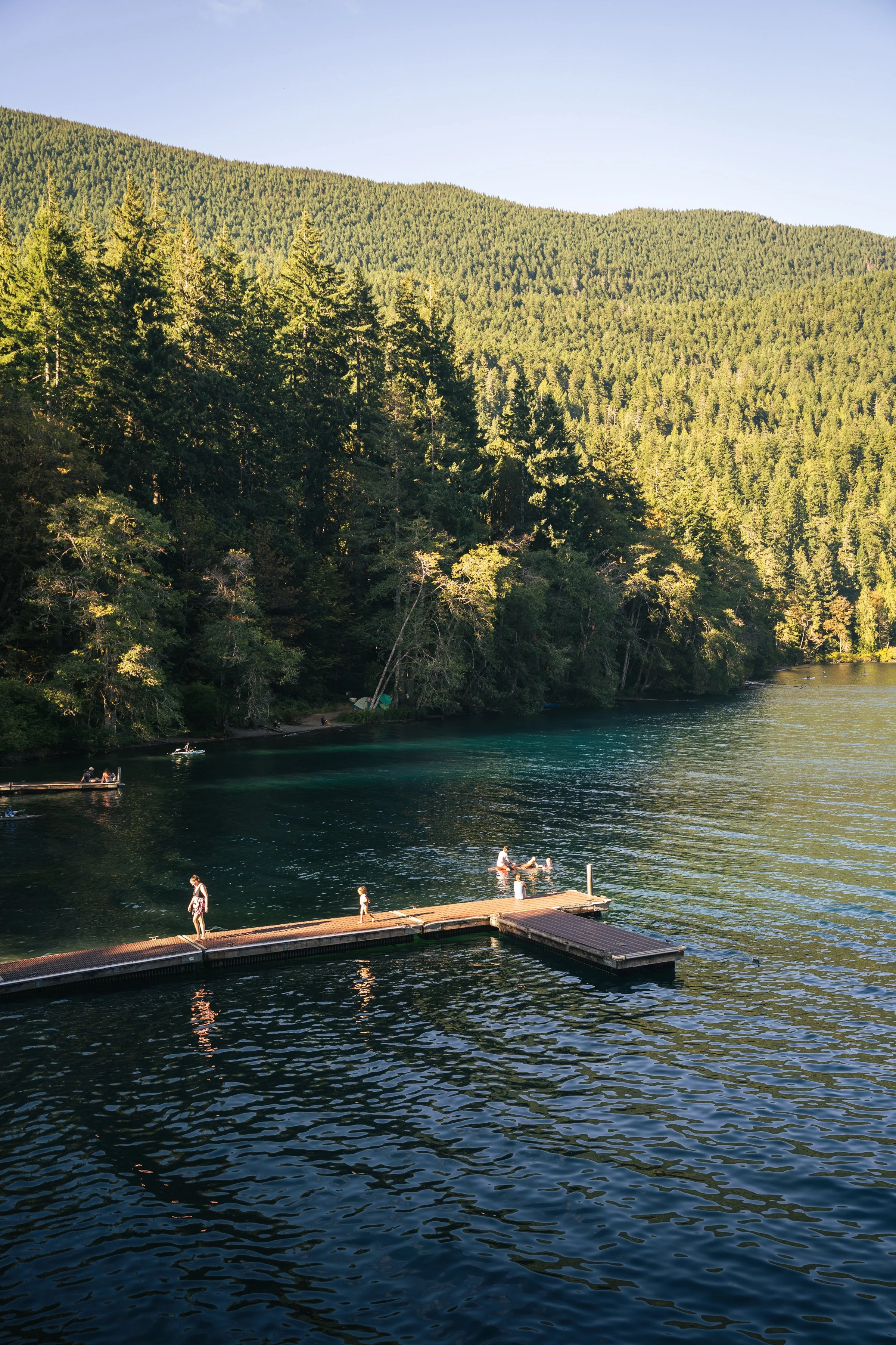 People enjoying a sunny day at a dock on a lake surrounded by lush green mountains and dense forest.
