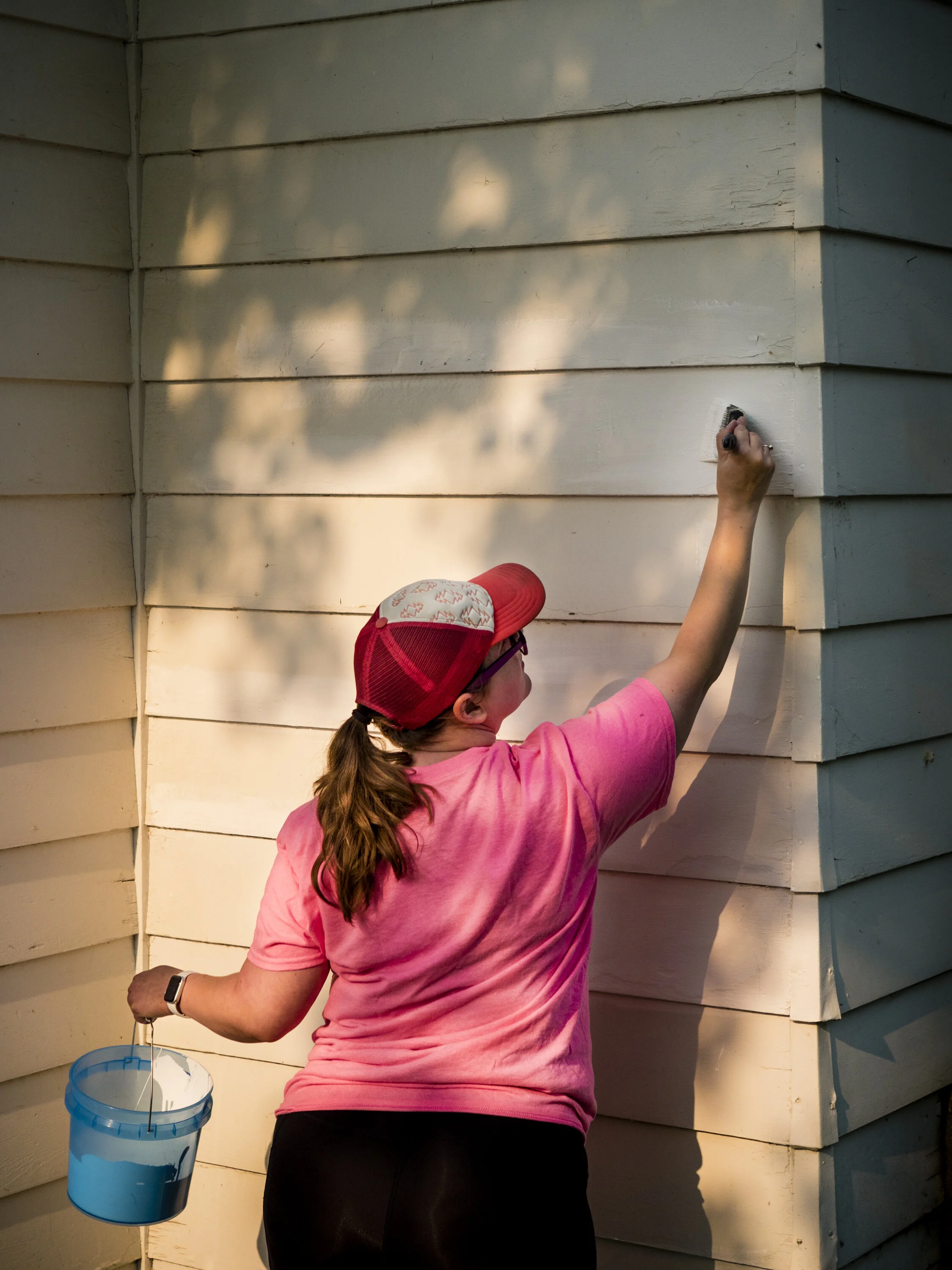 A person cleaning the exterior wall of a house with a brush, wearing a pink shirt, black pants, red and white cap, and a smartwatch on their left wrist. There is a blue bucket hanging on their arm.