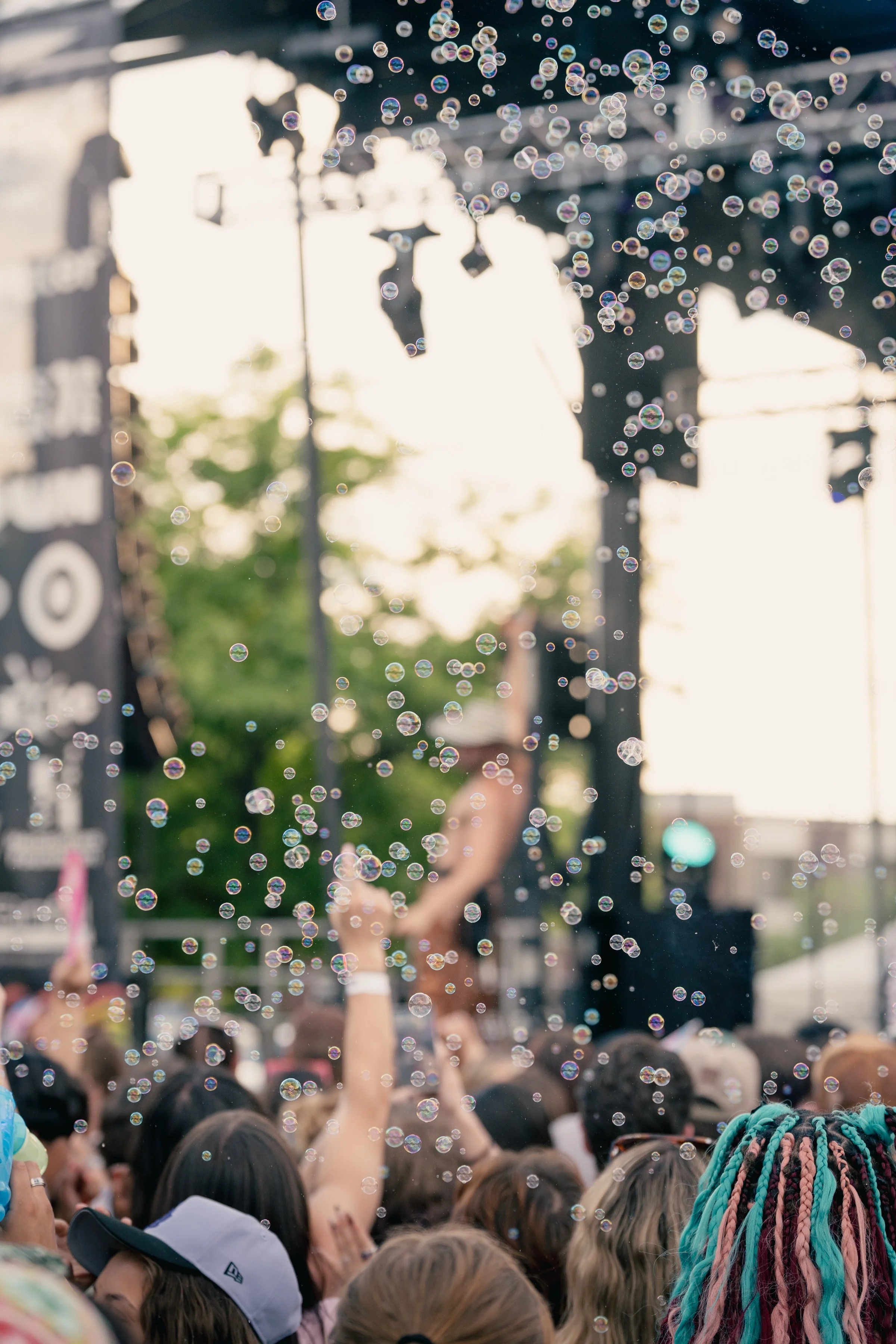 Crowd gathered outdoors at sunset with bubbles floating in the air, colorful braided hair, and a person holding up a phone to record, in a lively festival or concert setting.