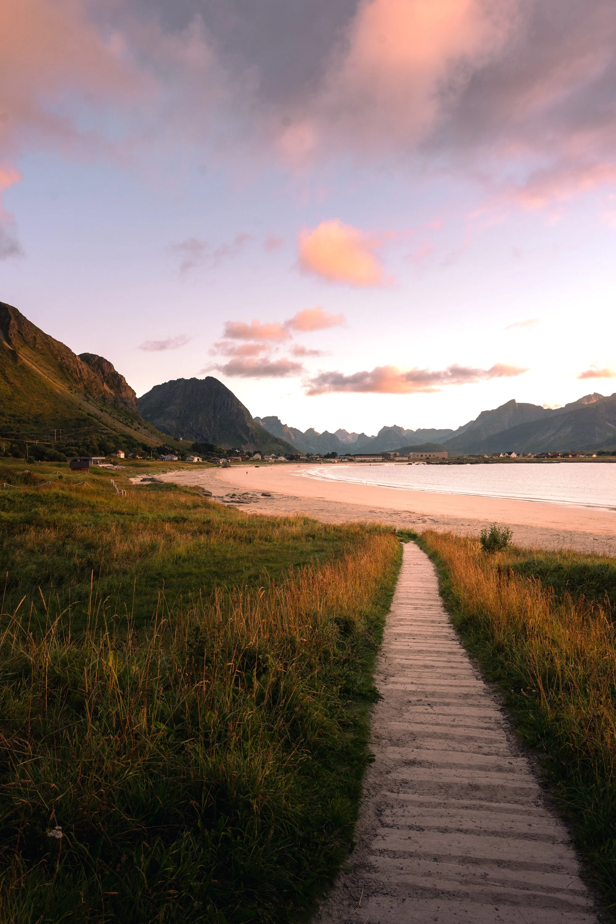 A scenic view of a sandy beach with a wooden pathway leading toward the water, surrounded by green grass and mountains in the distance under a sky with pink and purple clouds during sunset.