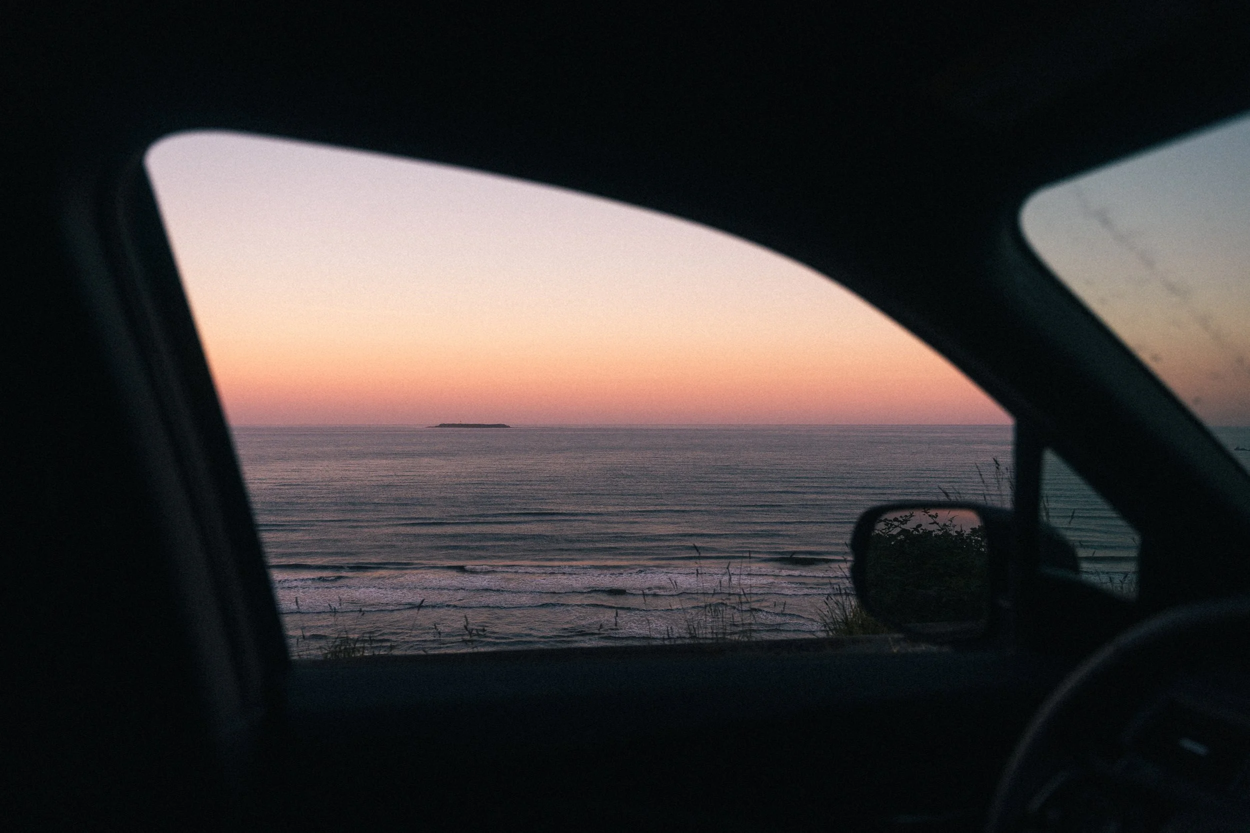 View of the ocean and a small island at sunset, seen through the windshield and side window of a vehicle.