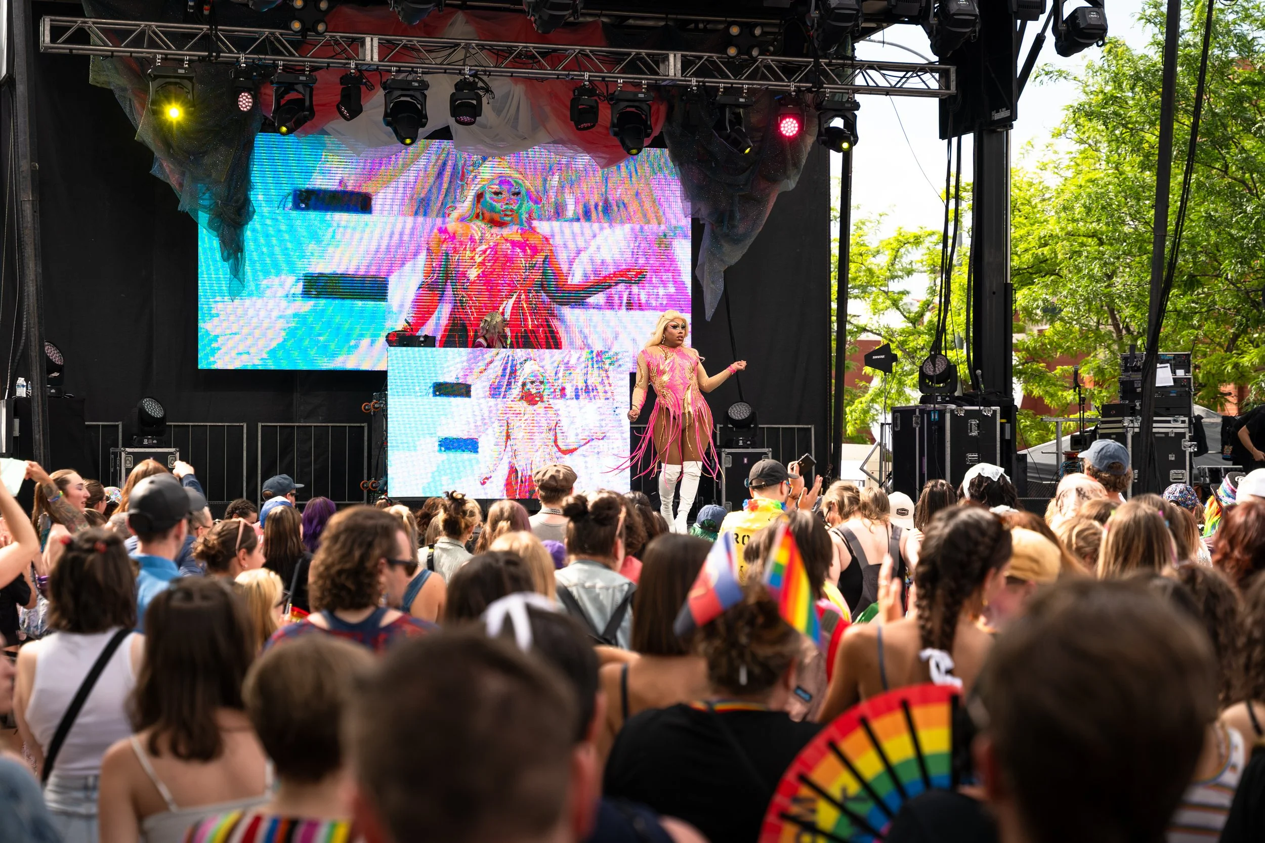 A stage with performers and a large digital screen displaying a colorful cartoonish face, set outdoors with trees and a crowd watching.