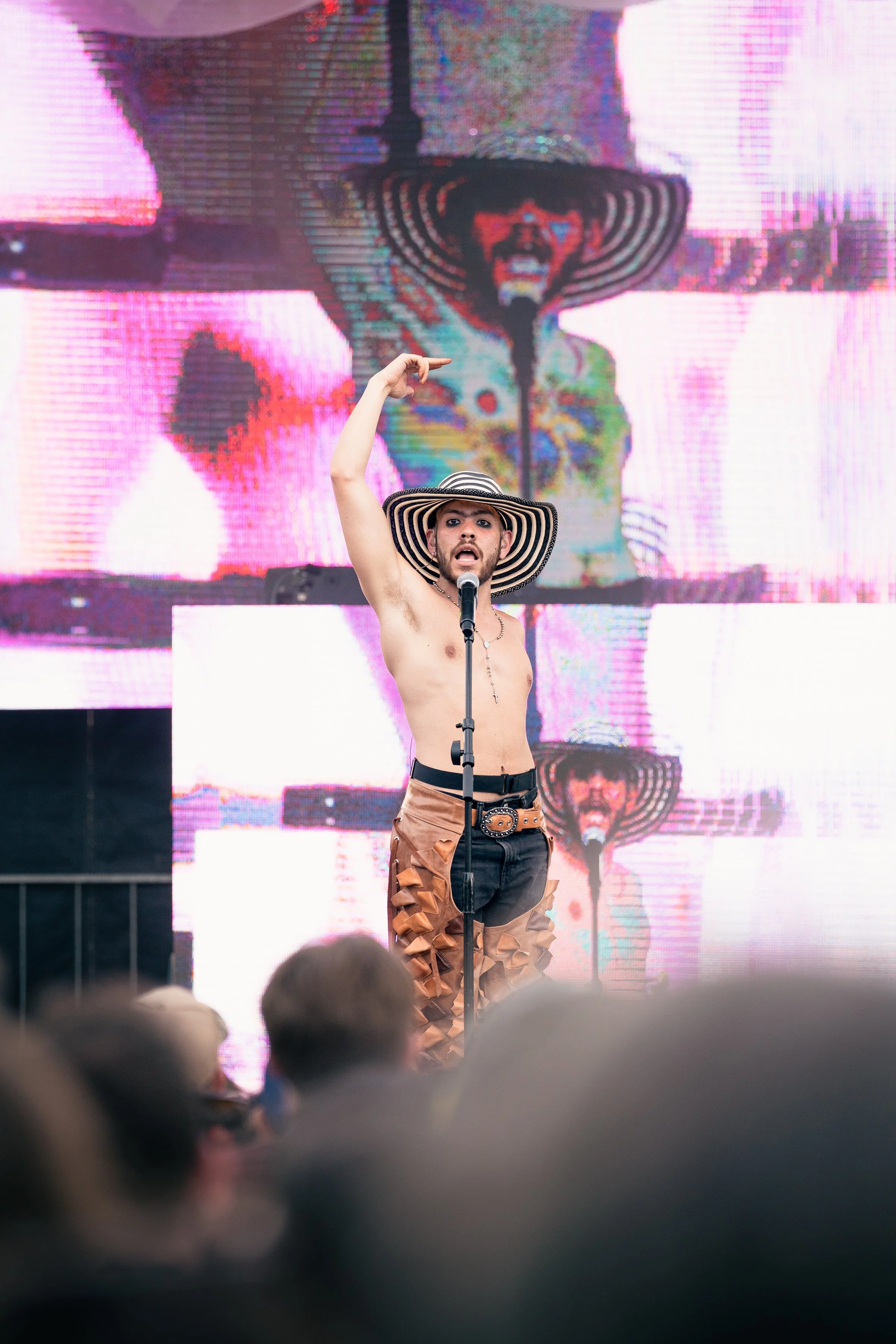 A shirtless man with a striped wide-brimmed hat standing in front of a large digital screen showing their own image. The man is pointing upward with one hand and has a microphone in front of him. The audience is visible at the bottom of the image.