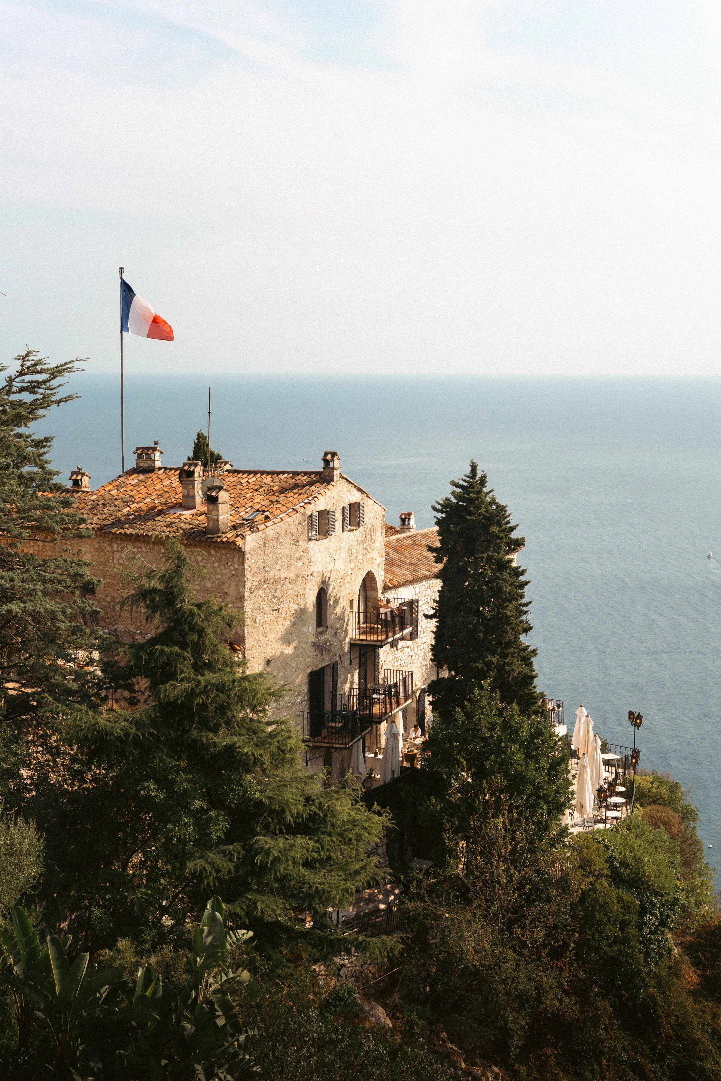 Cliffside restaurant with outdoor seating, surrounded by lush trees, overlooking the sea under a clear sky, with a building flying the French flag.