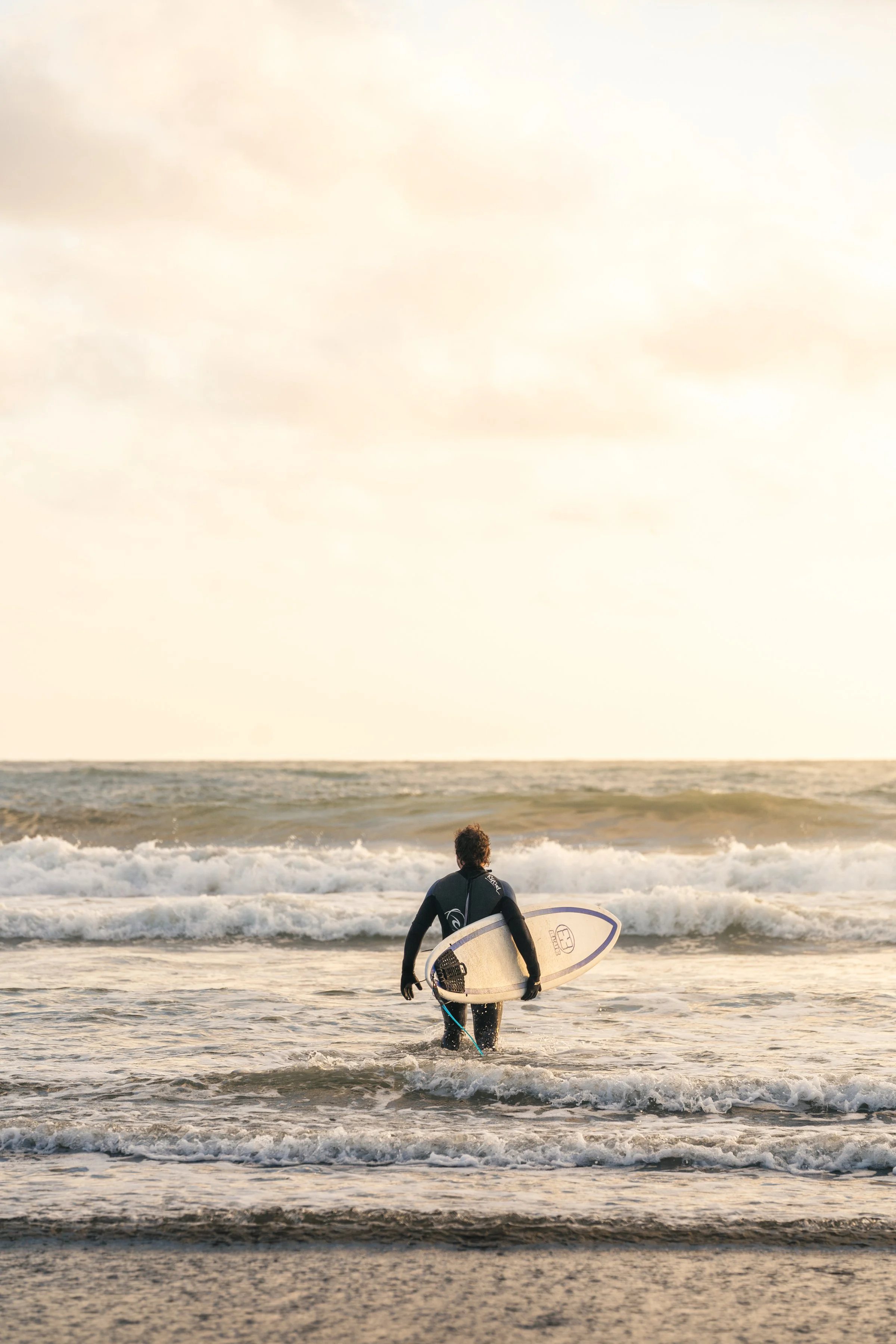 A person dressed in a black wetsuit holding a surfboard walking into the ocean at sunset.