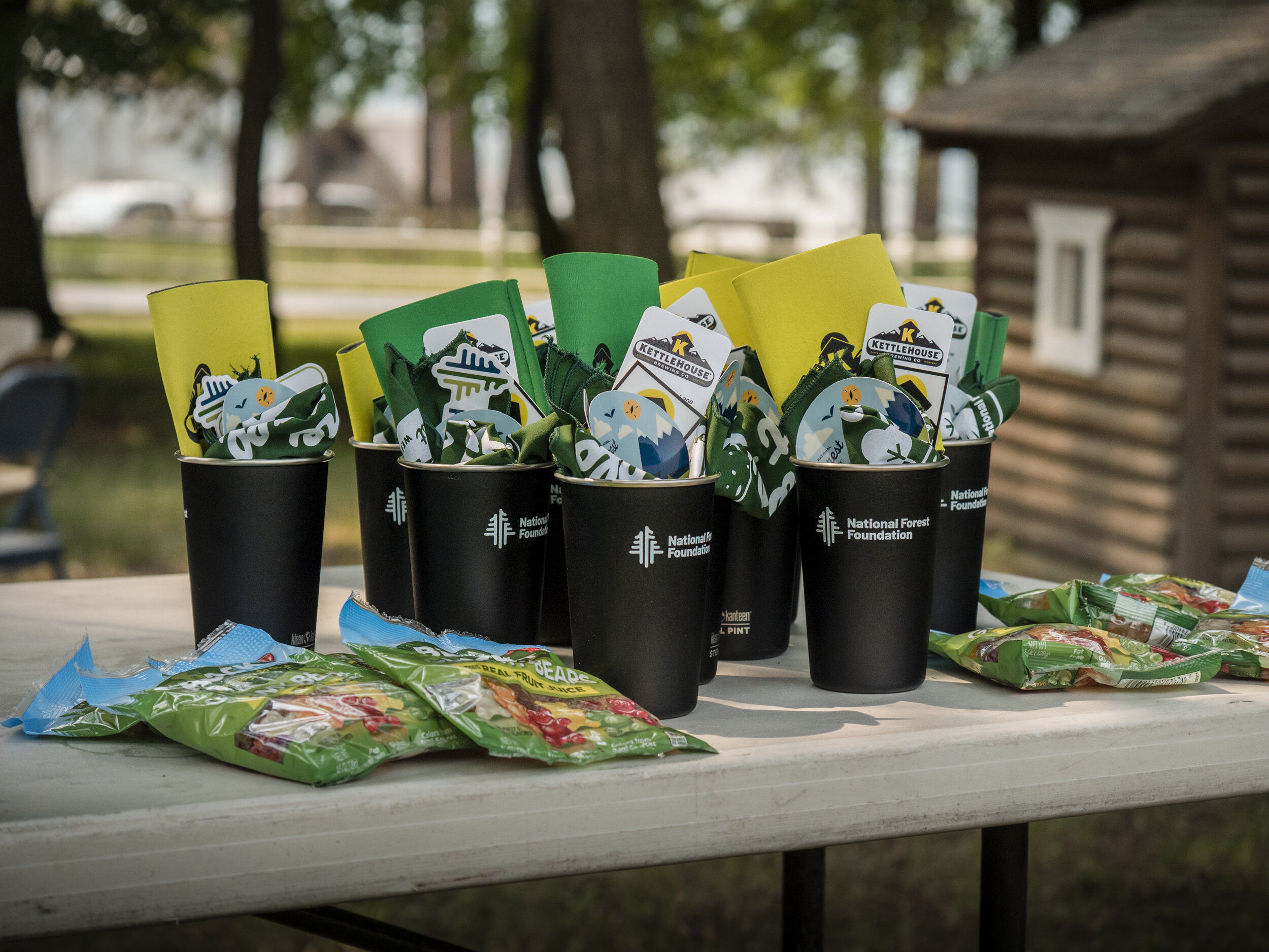 Black cups with logos and items inside, arranged on a white table outdoors. Surrounding trees and an outbuilding are visible in the background.
