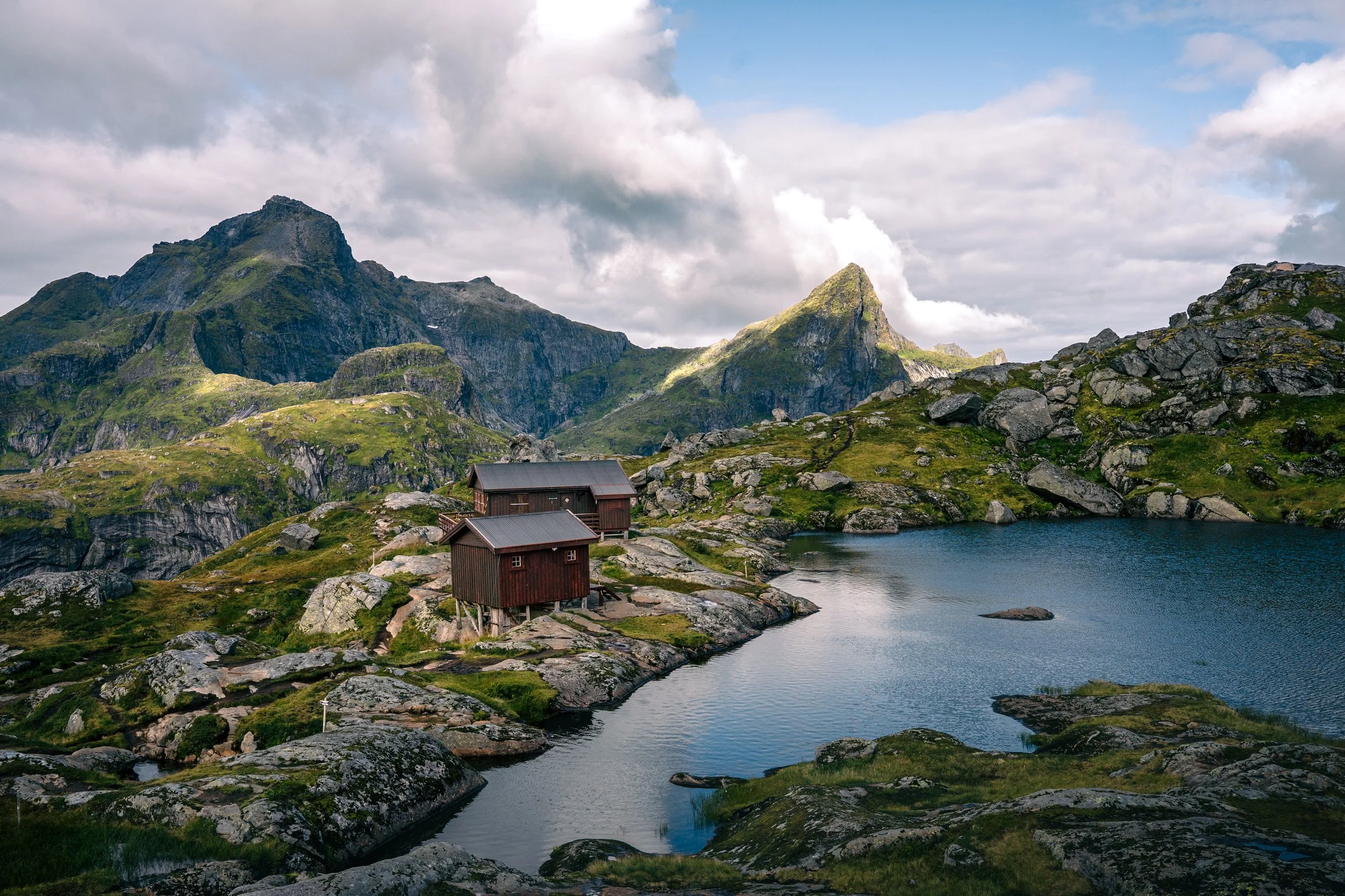 Scenic mountain landscape with green peaks, a calm lake, and a few wooden houses by the water under a cloudy sky.