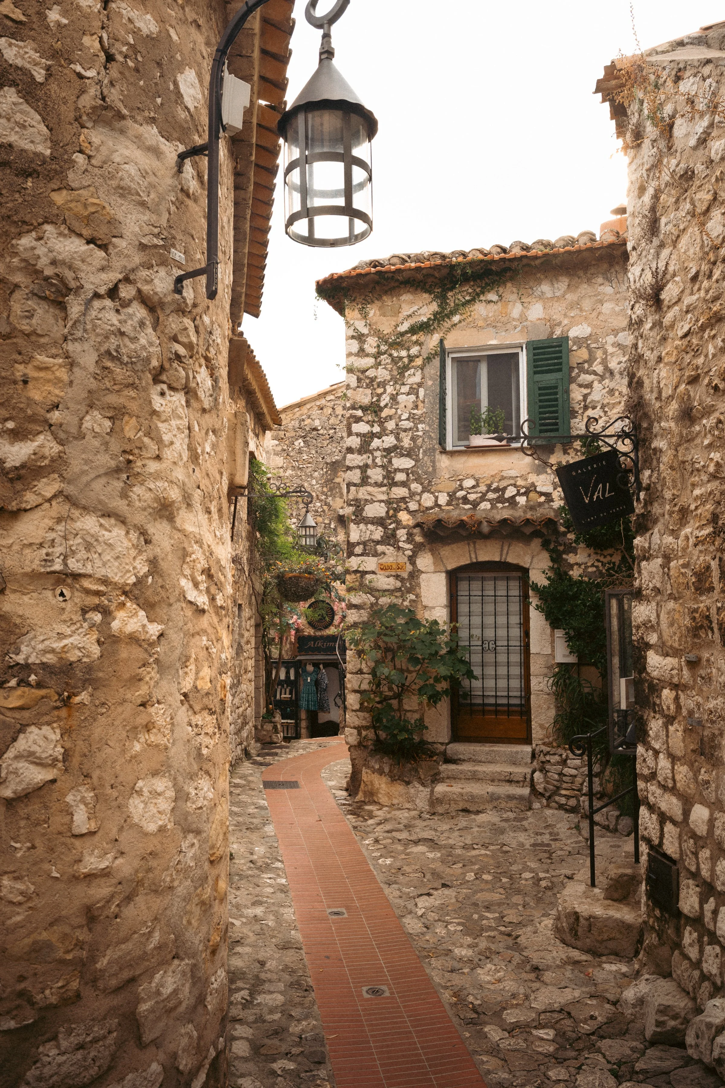 A narrow cobblestone alleyway in a rustic European village with stone buildings, a small tree, and hanging lanterns.