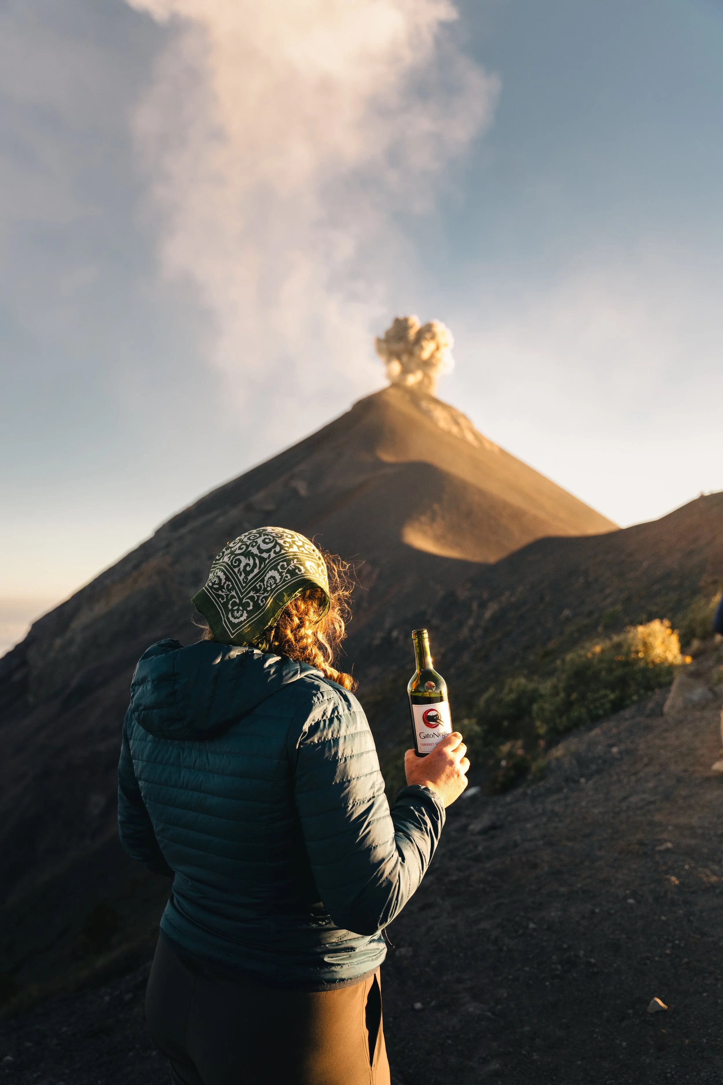 A person with a patterned bandana on their head, wearing a puffer jacket, holds a bottle of wine with a mountain in the background, emitting steam or smoke, possibly a volcano.