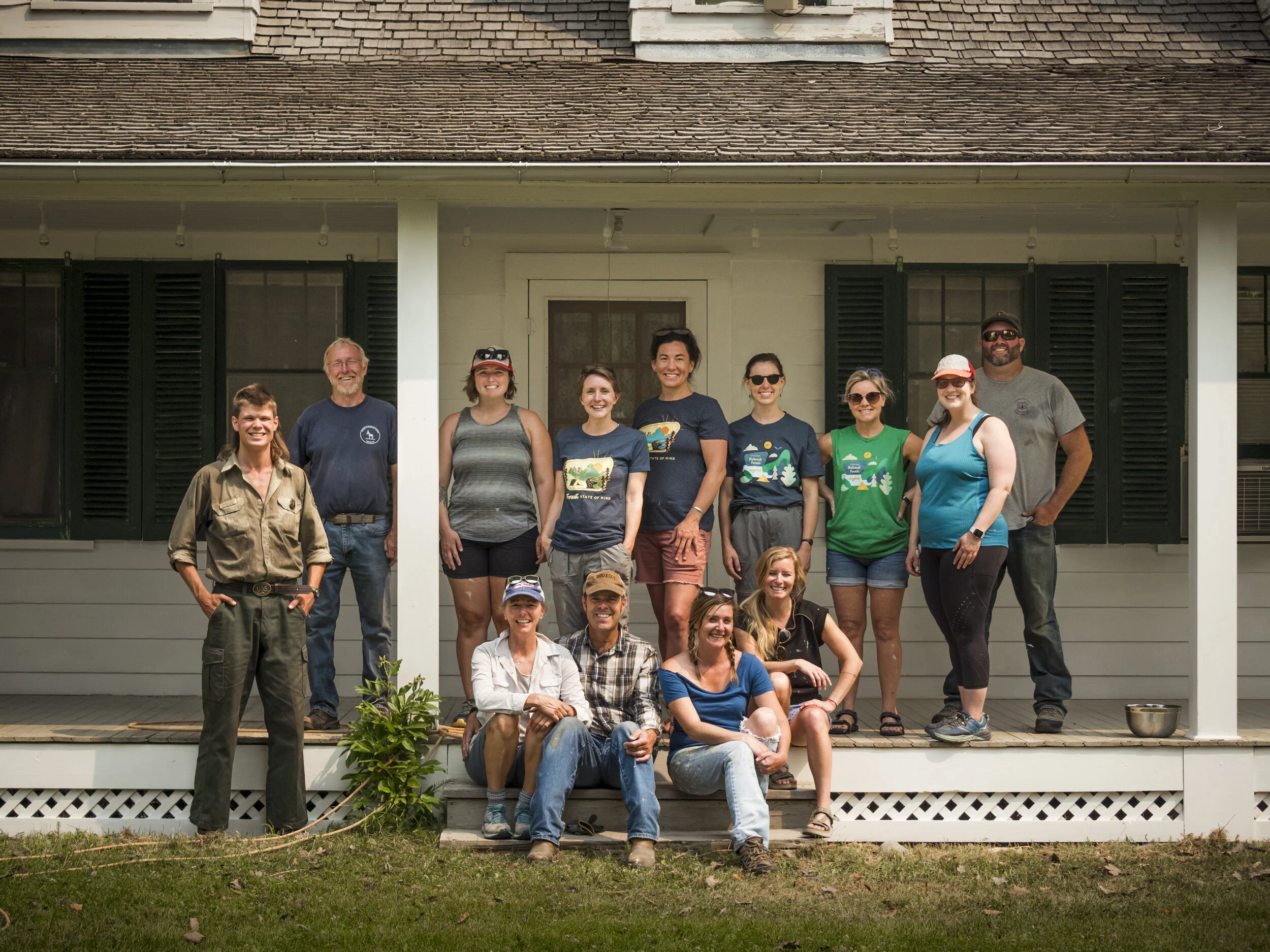 A group of ten people standing on the porch of a house, smiling at the camera. The house is painted light gray with black shutters and has a shingled roof. The group includes men and women, some wearing casual clothes, and appears to be enjoying a ga