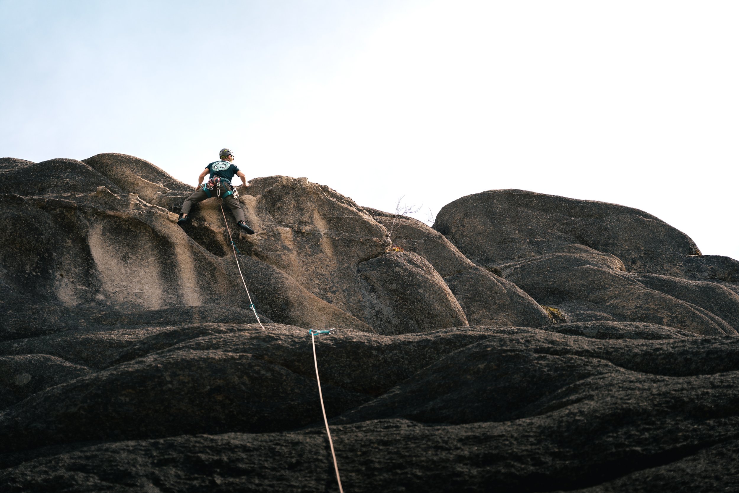 A person rock climbing on a large, dark rock formation outdoors, wearing climbing gear and a helmet, with a climbing rope attached.